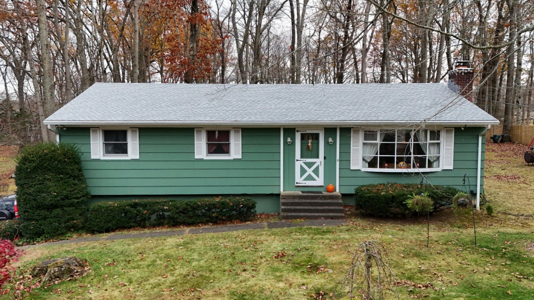 Green ranch house with white trim, a small front porch, and a brown brick chimney against a backdrop of trees.