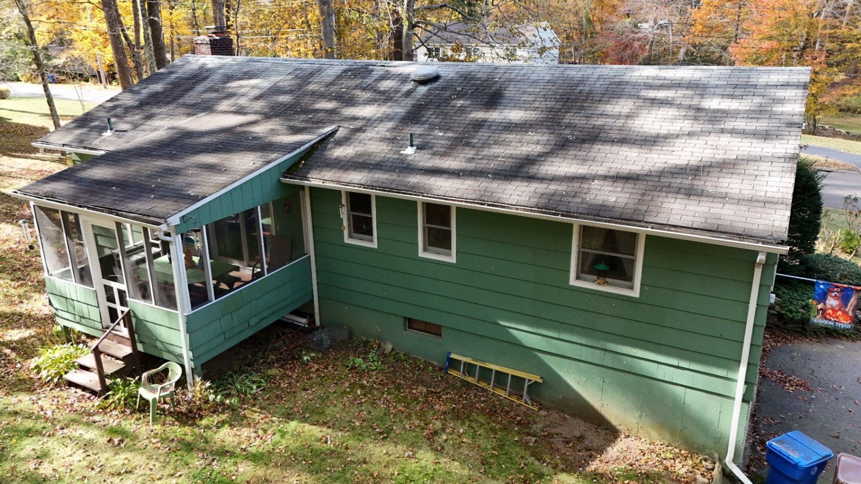 Green house with attached screened porch and sloping roof; fall foliage in the background.