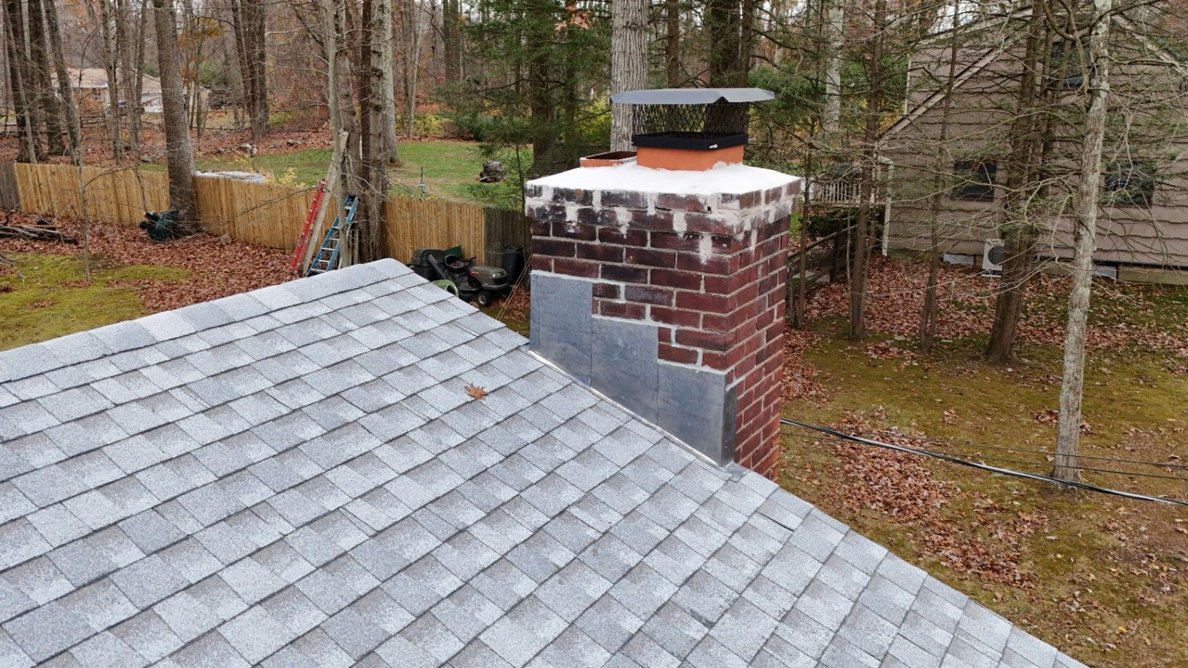Brick chimney with a metal cap on a gray shingled roof, set in a wooded area.