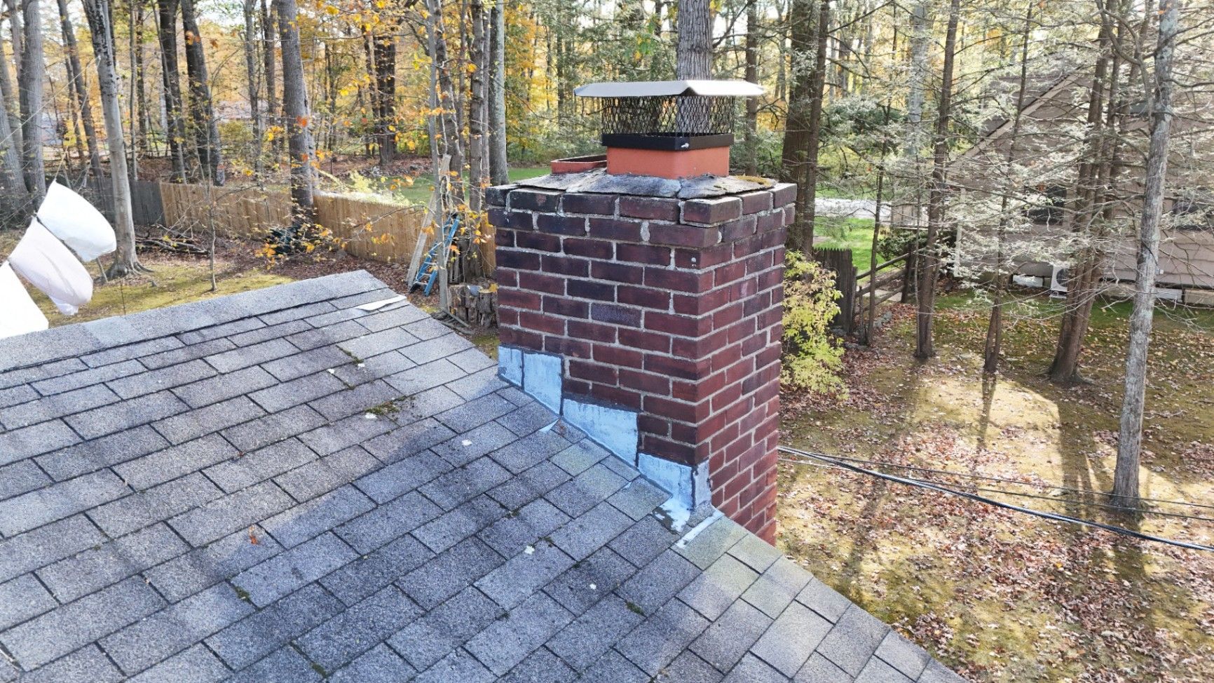 Brick chimney with flashing, on a shingled roof, surrounded by trees.