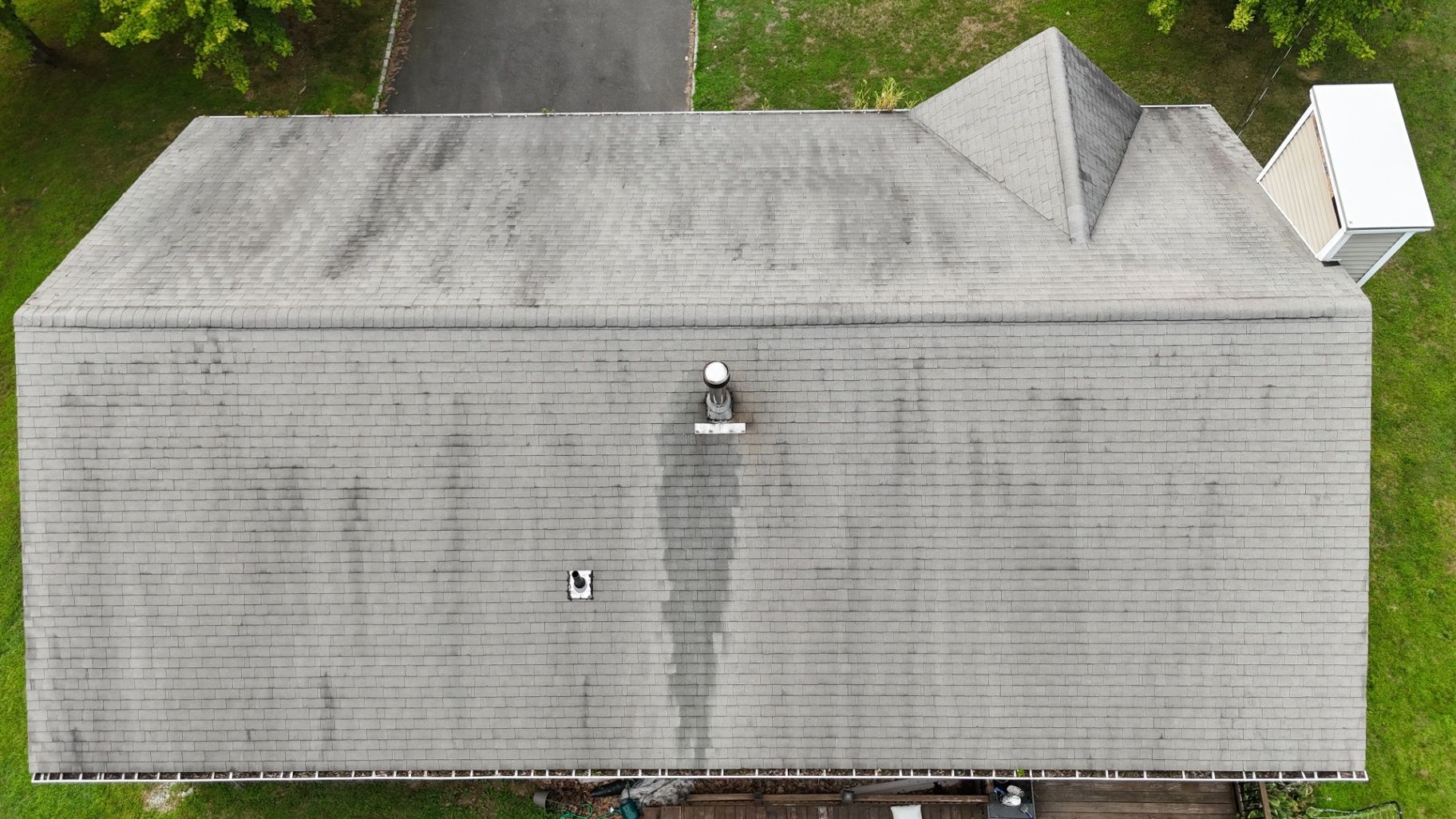 Overhead view of a gray asphalt shingle roof with a chimney and vent on a house, surrounded by green grass.