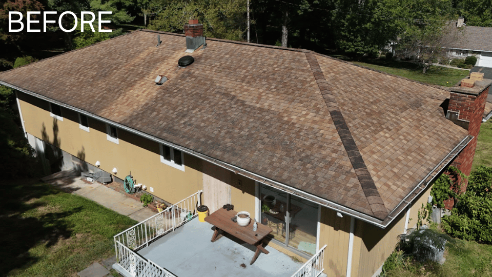 Aerial view of a brown-shingled roof on a yellow house before a cleaning. Green yard surrounds the building.