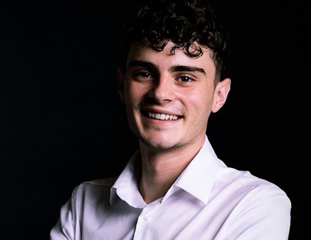 Ein junger Mann mit lockigem Haar trägt ein weißes Hemd und lächelt mit verschränkten Armen.