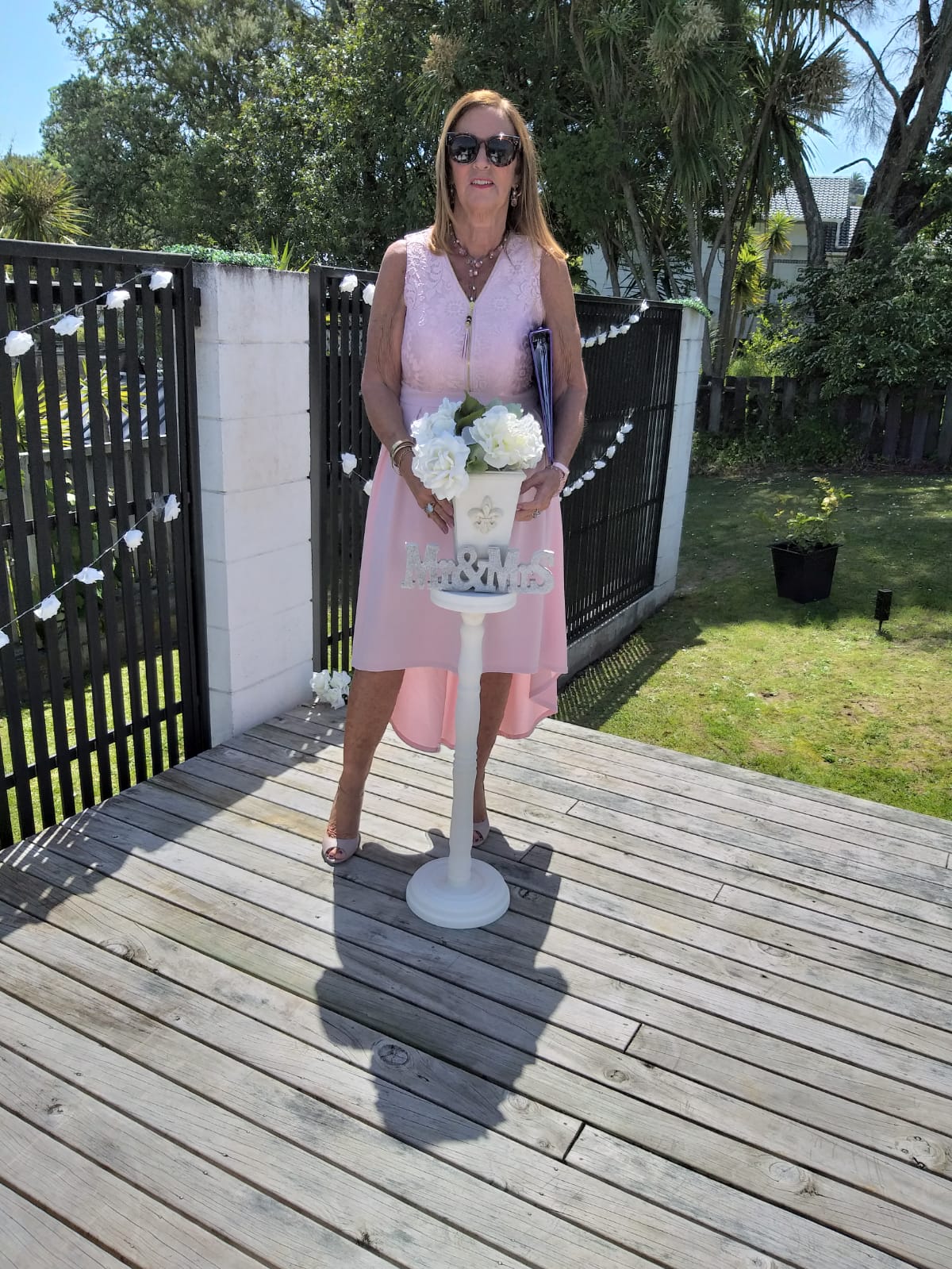 A woman in a pink dress is standing on a wooden deck holding a vase of flowers.