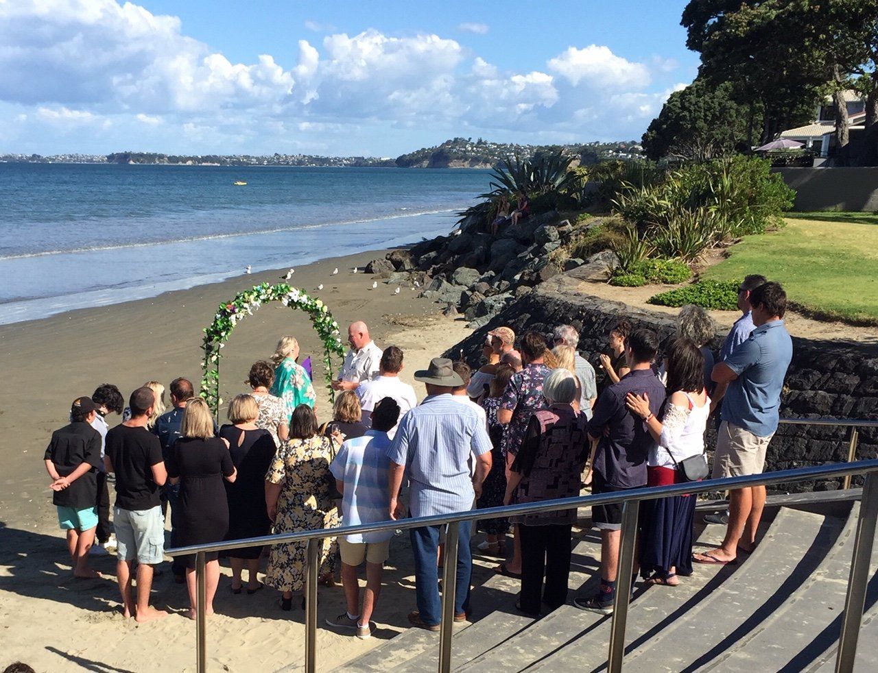 A group of people are standing on a beach watching a wedding ceremony.