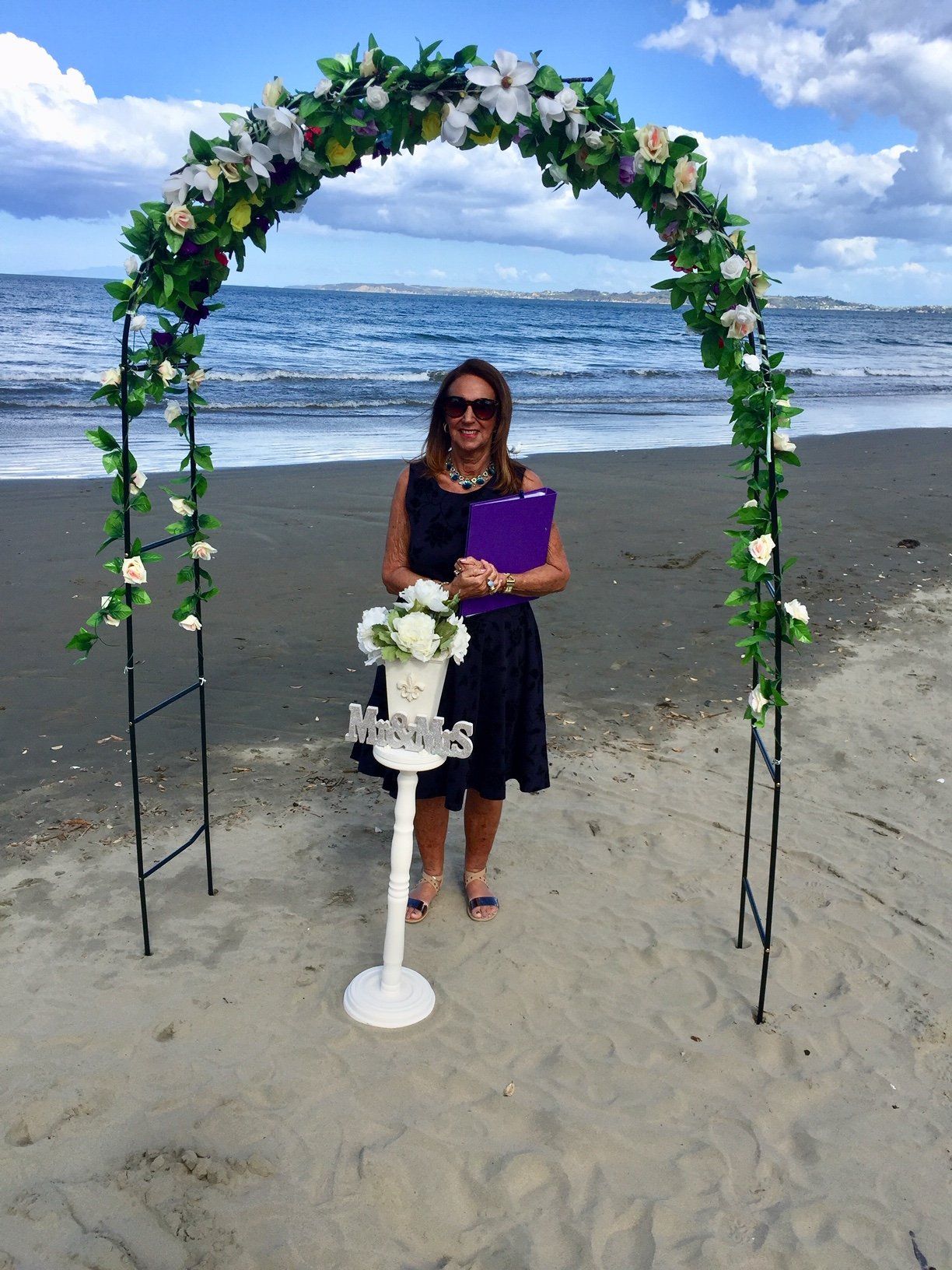 A woman in a black dress is standing in front of a floral arch on the beach.