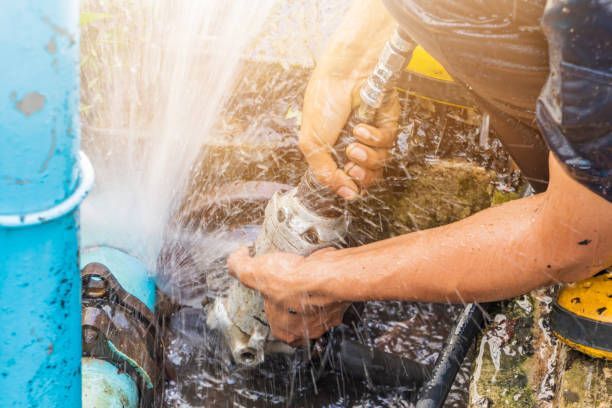 Person repairing a burst water pipe, water spraying out. Blue and black pipes, yellow shoe visible.