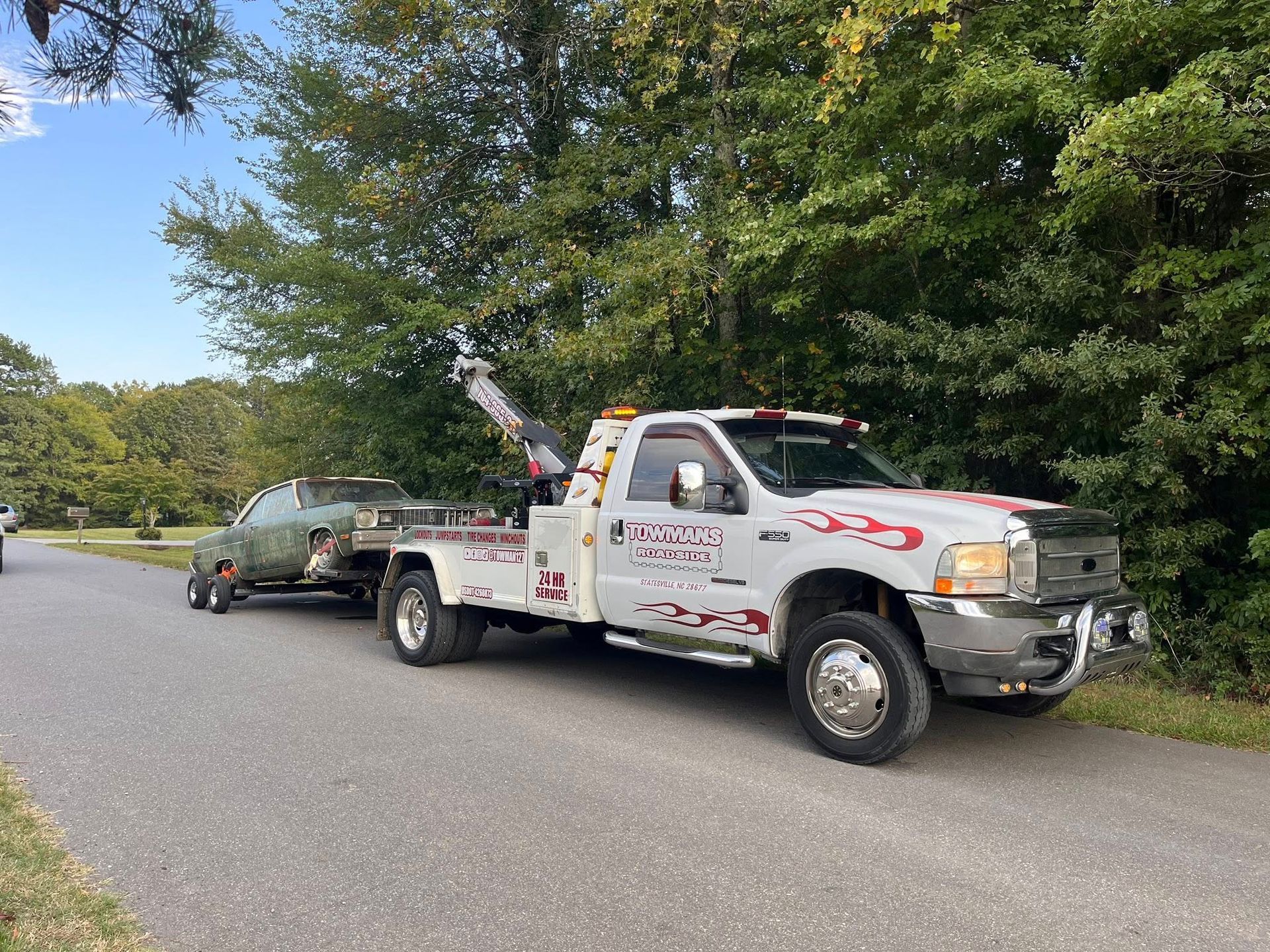 Tow truck hauling an old, green car on a tree-lined road. The truck has flames decals.