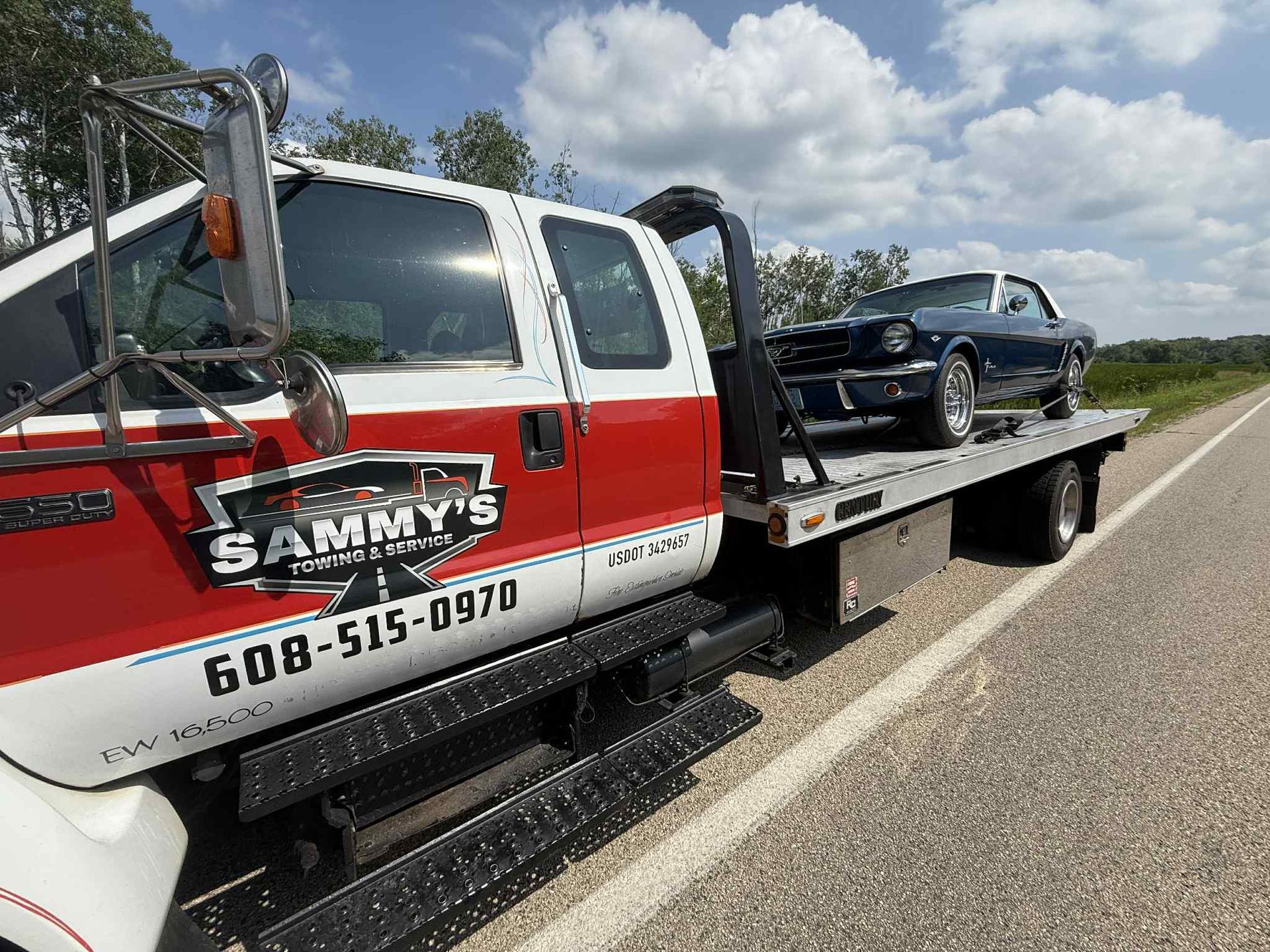 Tow truck with a classic blue car on its flatbed on a roadside under a cloudy sky.