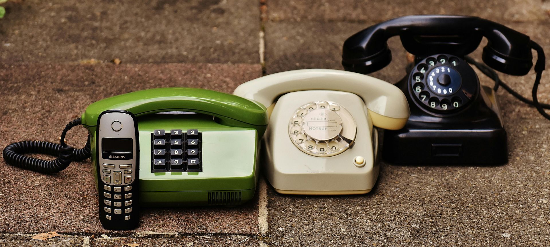 Several landline phones of varying styles and a cell phone, all resting on a stone surface.