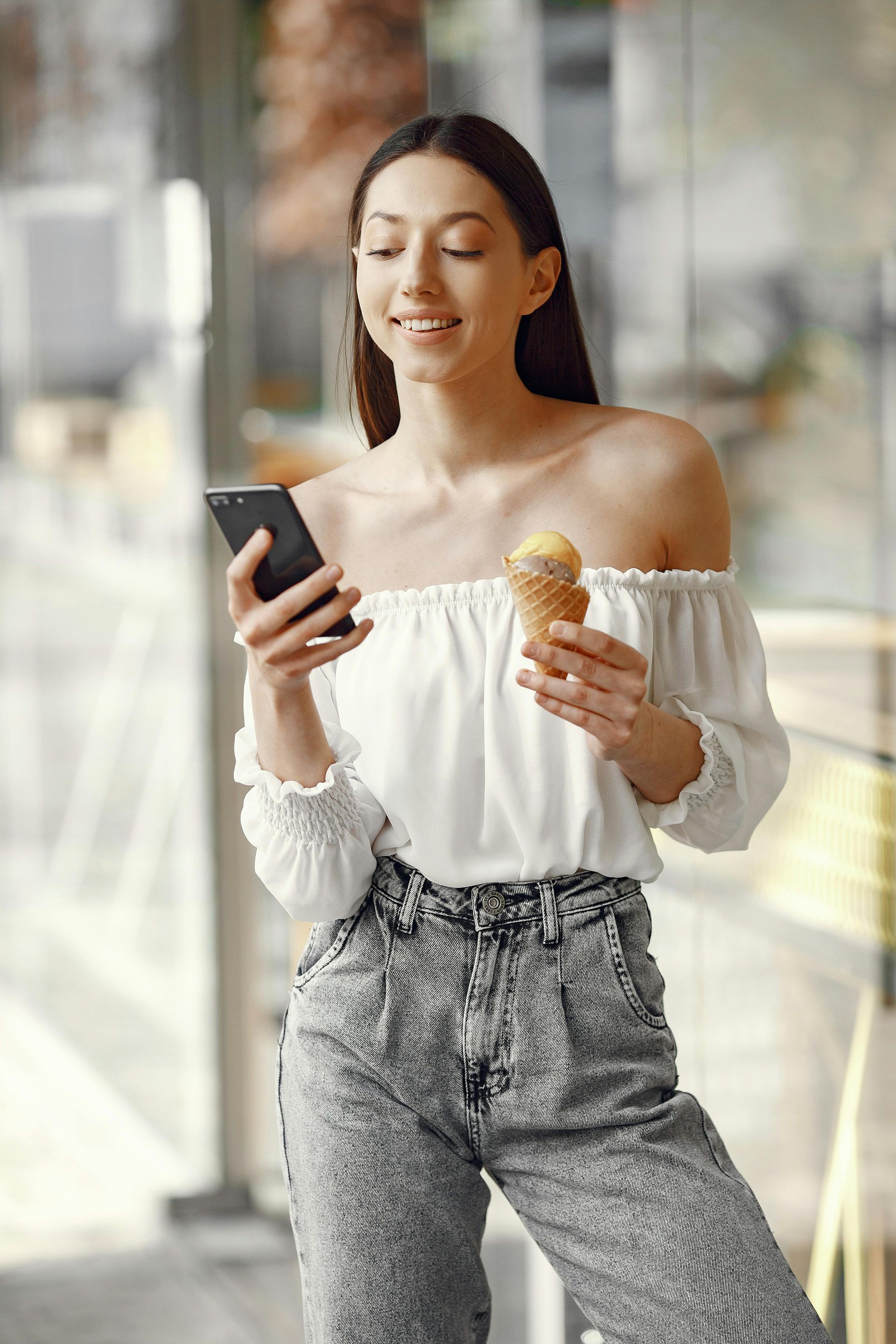 A person with long brown hair, wearing a white off-shoulder top and patterned pants, holds a smartphone and ice cream.