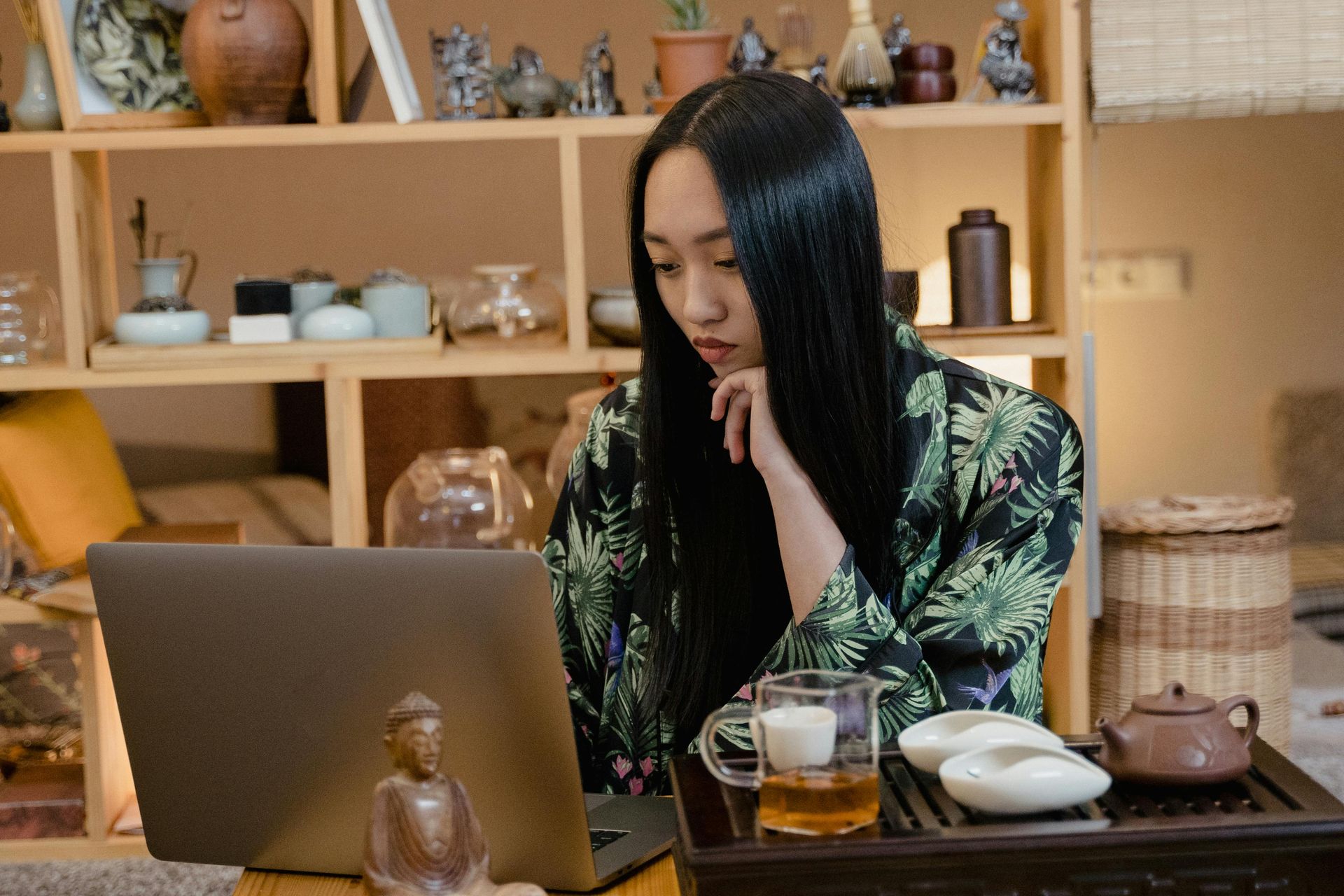 Woman with long dark hair, wearing a green kimono, looking at laptop, tea set nearby, sitting in front of a shelf.