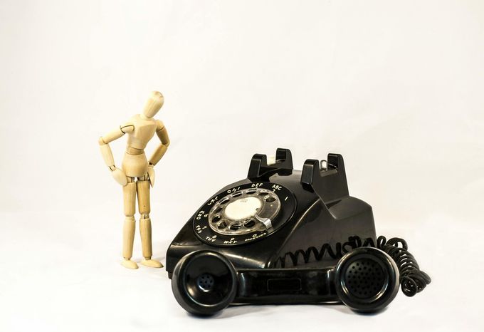 Wooden mannequin looking at a black rotary phone on a white background.