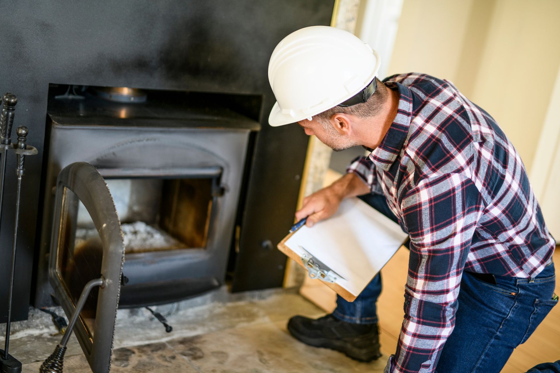 Man in hard hat examines a wood-burning stove, holding clipboard, assessing inside the fireplace.
