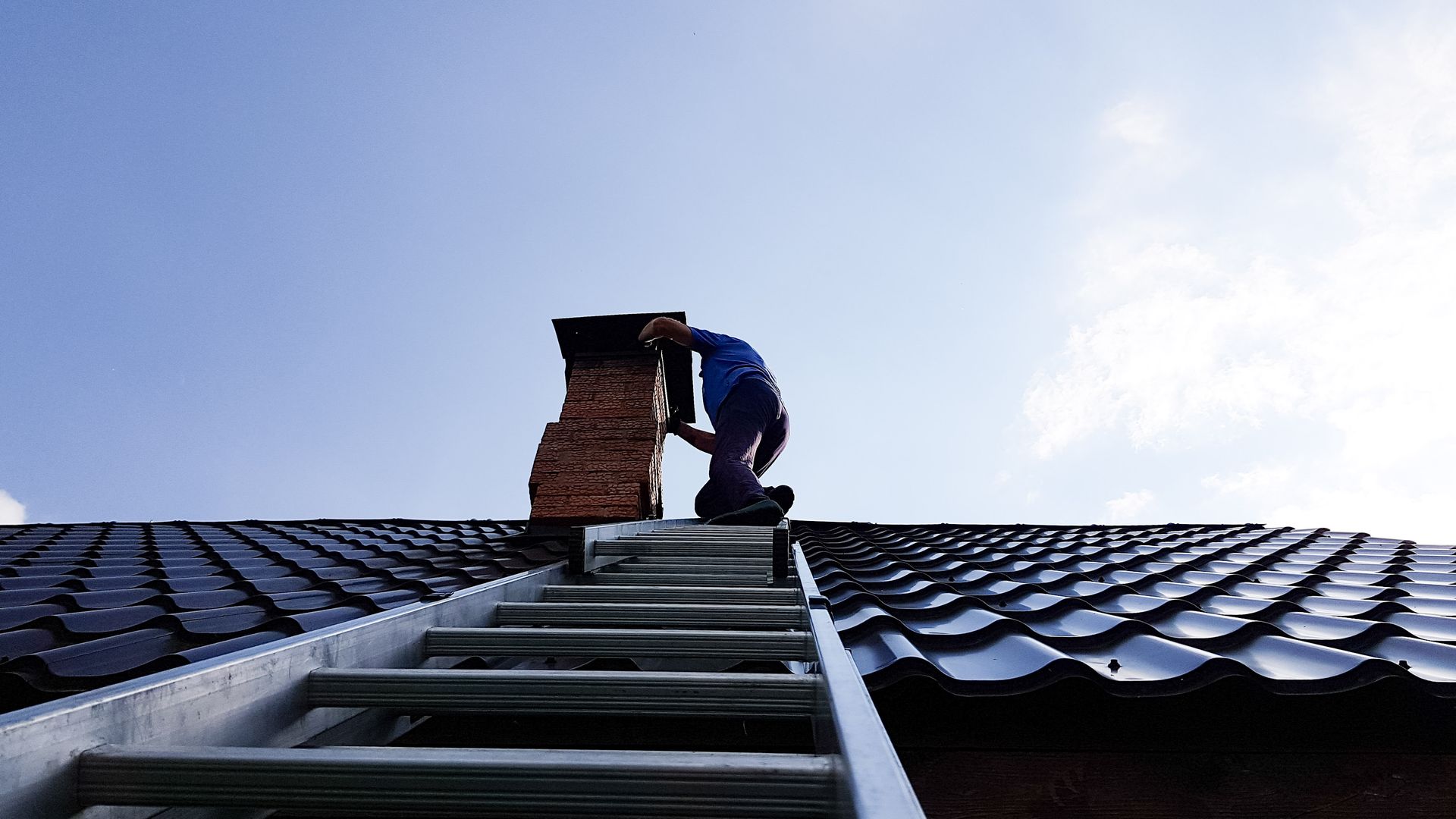 Person on a ladder, cleaning a chimney on a rooftop under a blue sky.