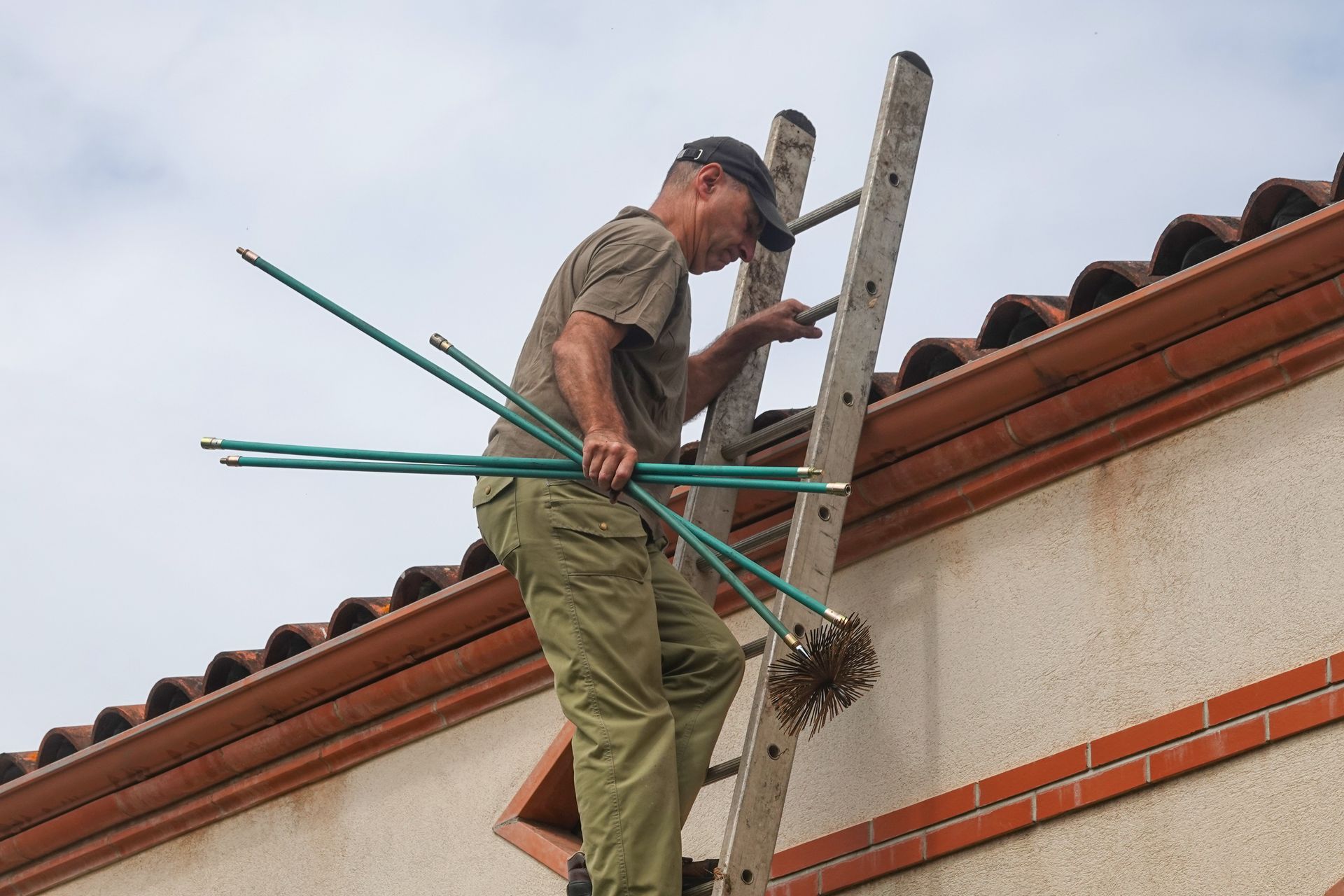 Man climbing ladder onto a roof, holding chimney cleaning rods and brush.