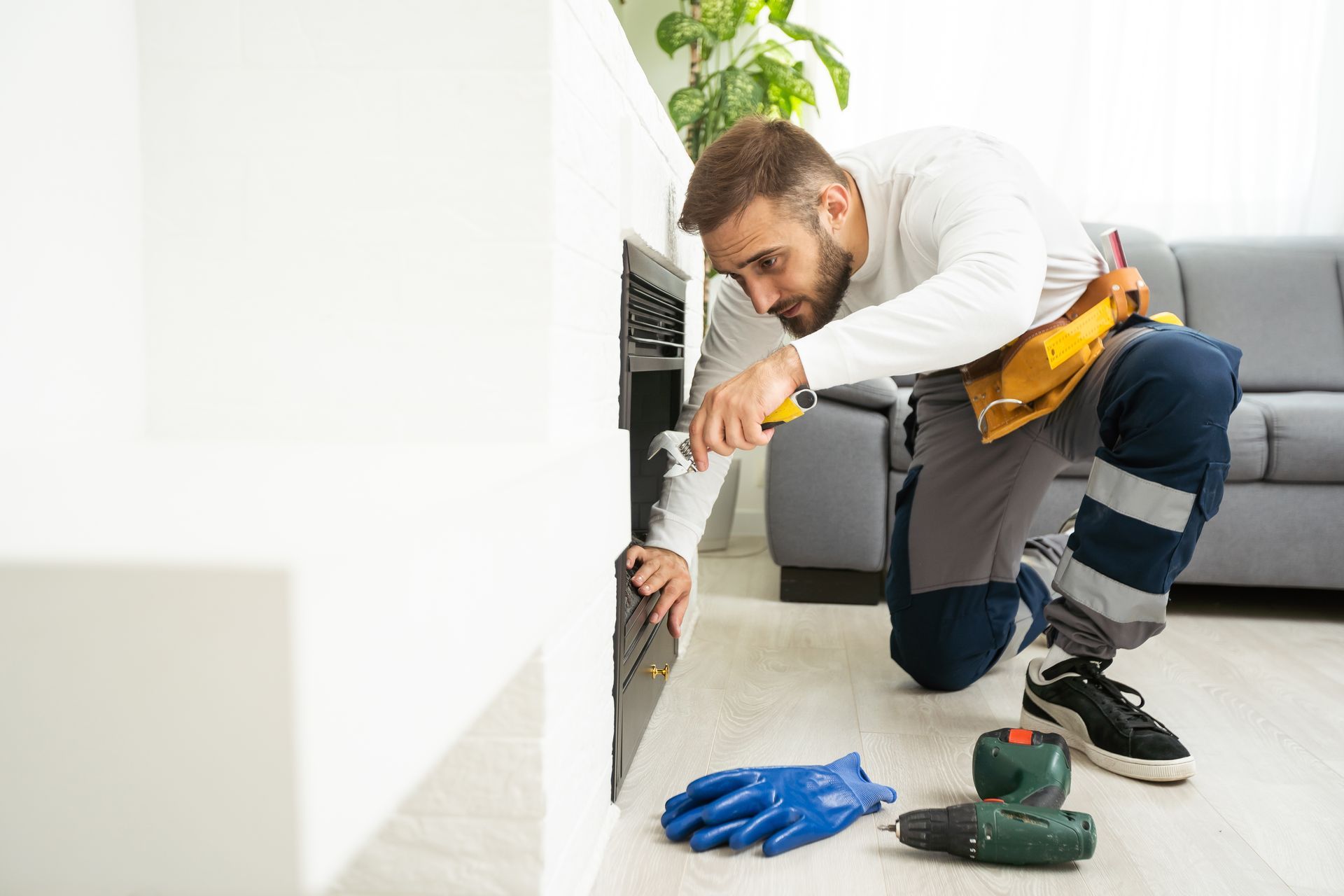 Man in work clothes kneeling, using a tool on a dark panel in a room. Tools and gloves lie nearby.