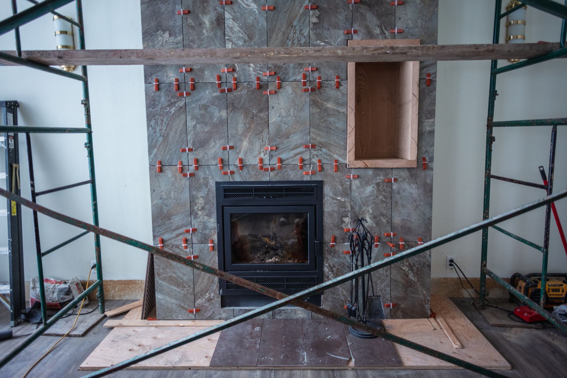 Fireplace under construction, covered in gray tile, with orange tile spacers and wooden scaffolding.