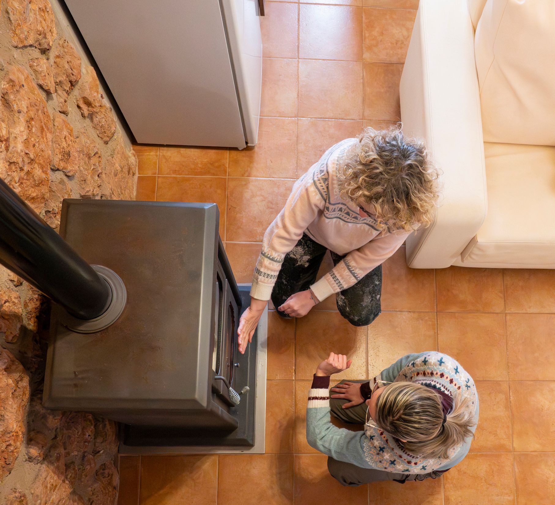 Two people kneeling by a dark stove, one touching it; tiled floor, stone wall, white couch.