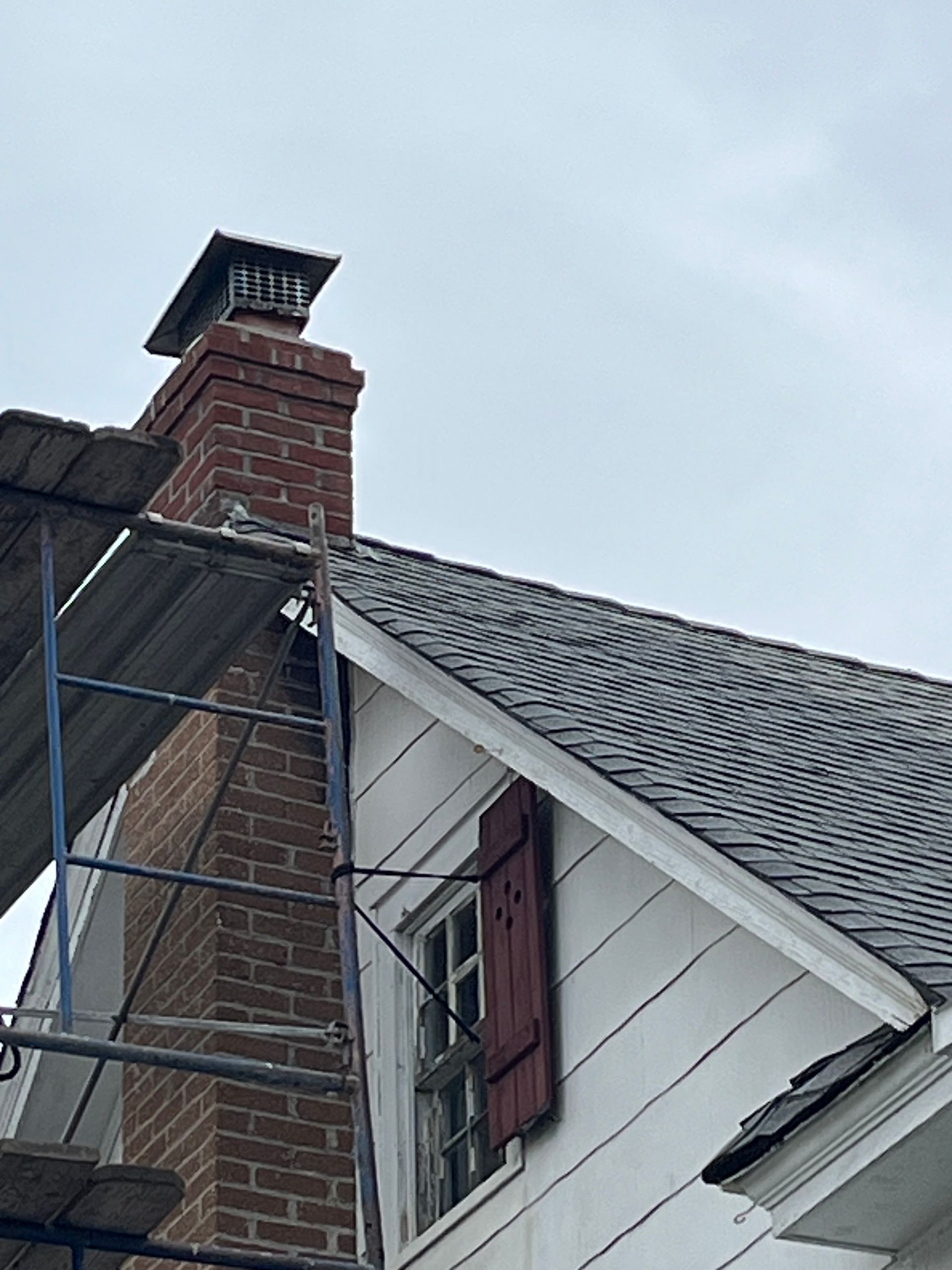 Chimney on a house with scaffolding; red brick, black roof, white siding, and cloudy sky.