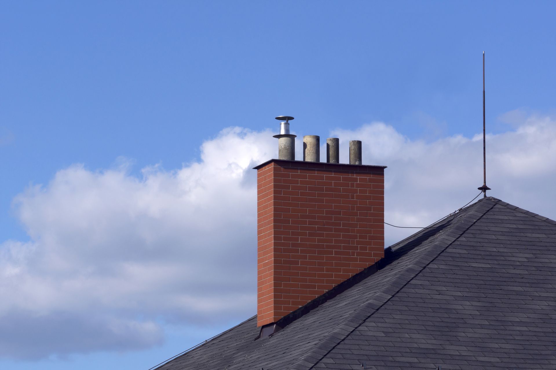 Brick chimney on a dark roof with a lightning rod, against a blue sky with fluffy white clouds.