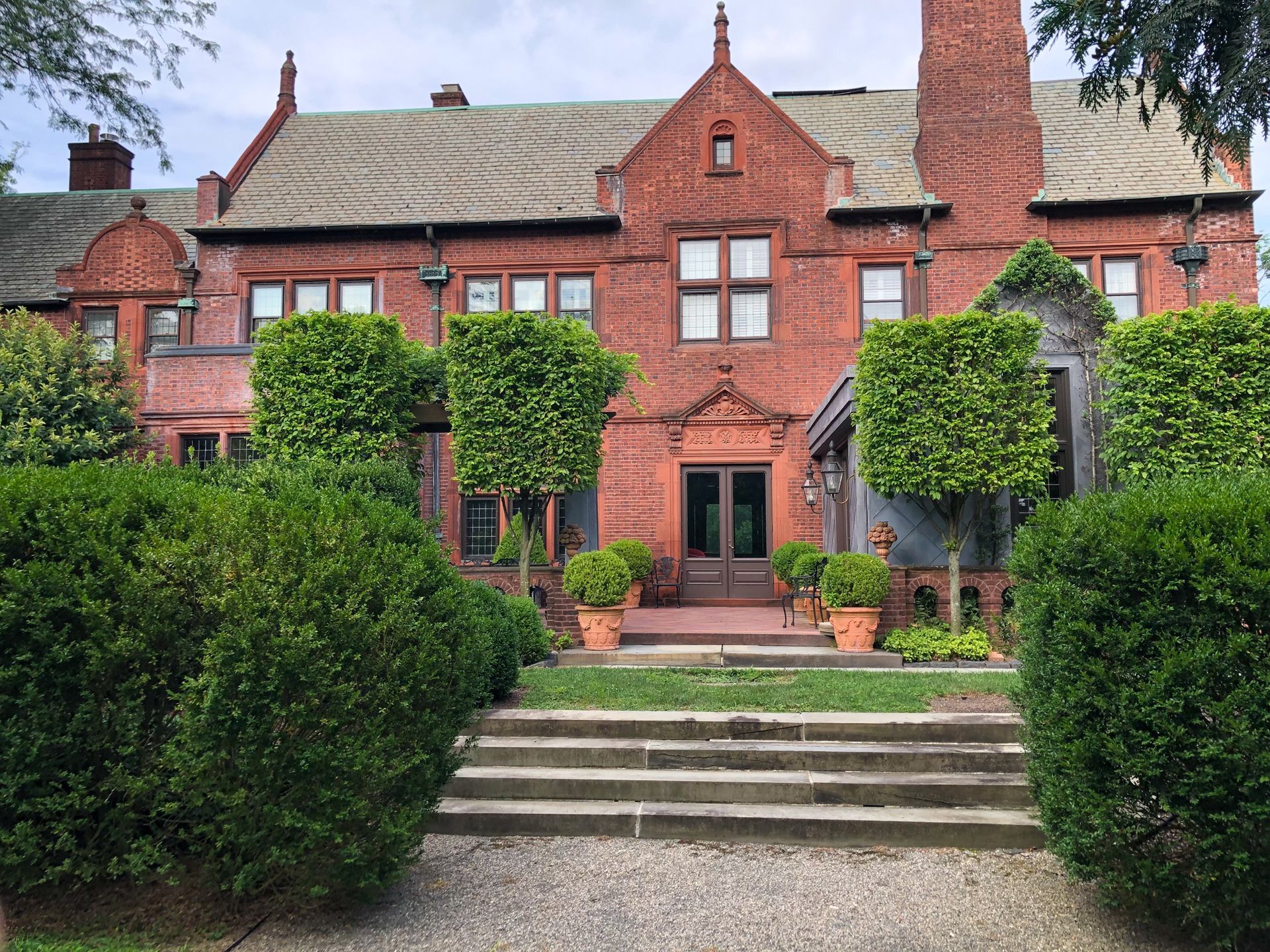 A large brick house with stairs leading up to it is surrounded by bushes and trees.