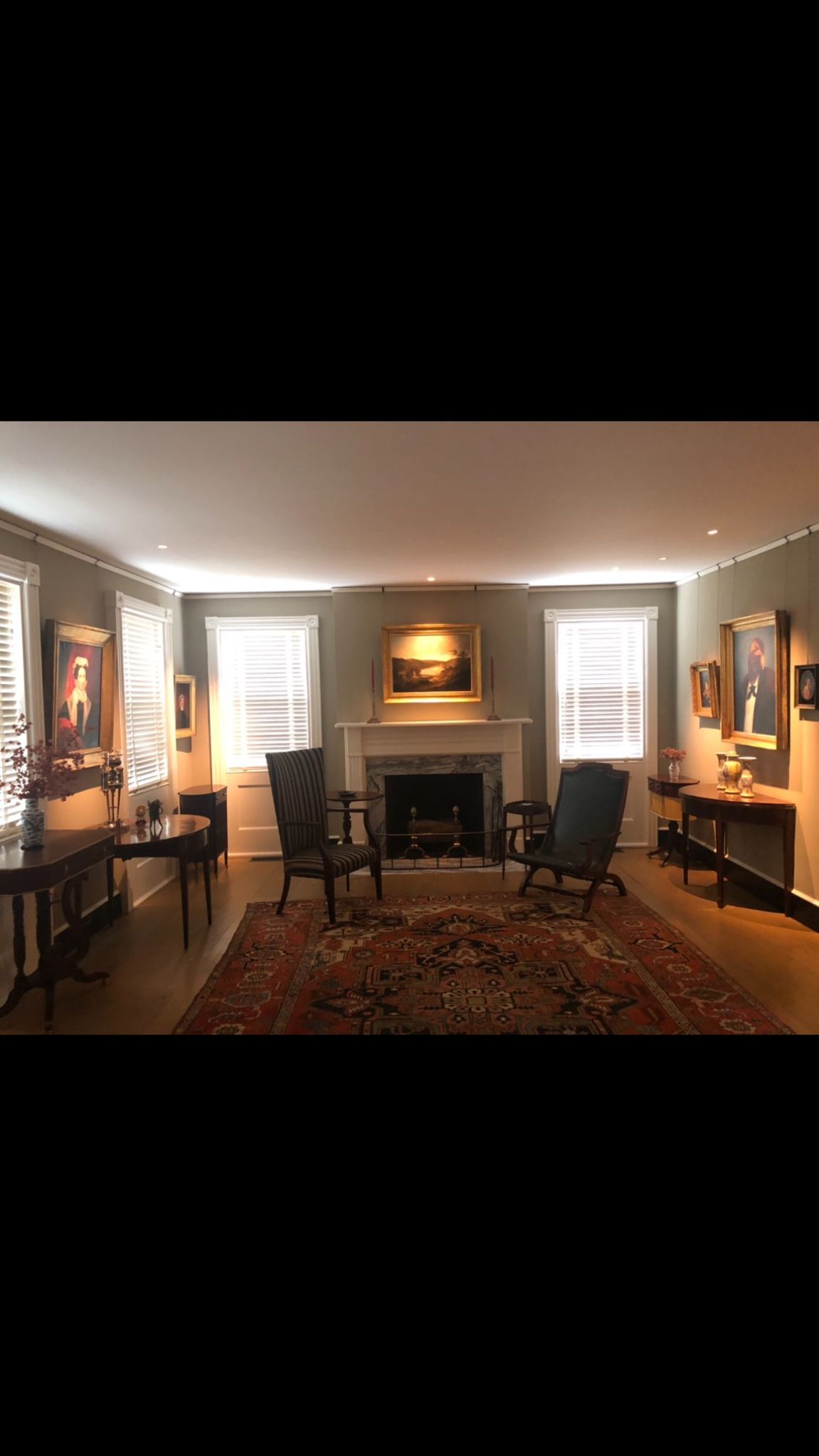 A living room with a fireplace , chairs , tables and paintings.