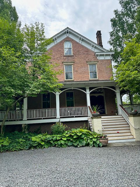 A large brick house with a porch and stairs is surrounded by trees.