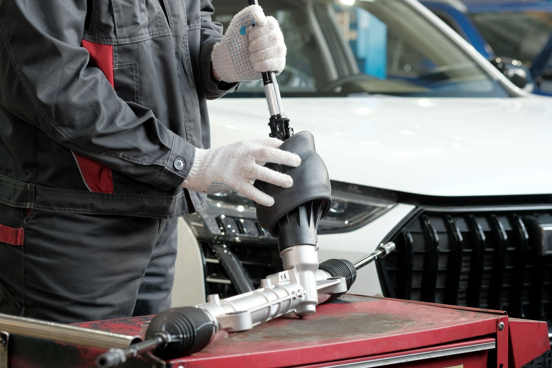 Mechanic in gray jumpsuit working on car steering rack in a shop.