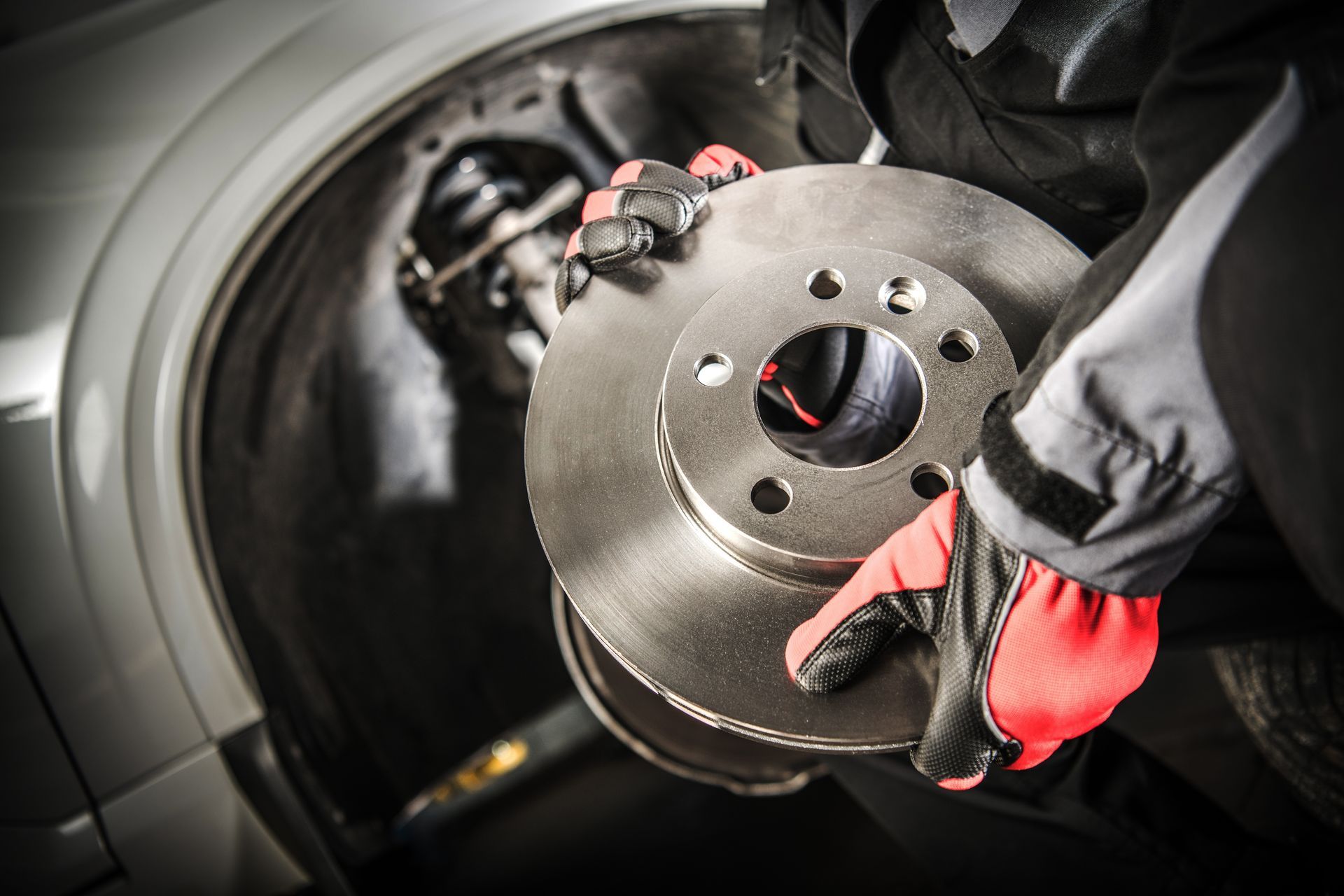 Mechanic holding a new car brake rotor, wearing red and black work gloves, near a vehicle's wheel well.