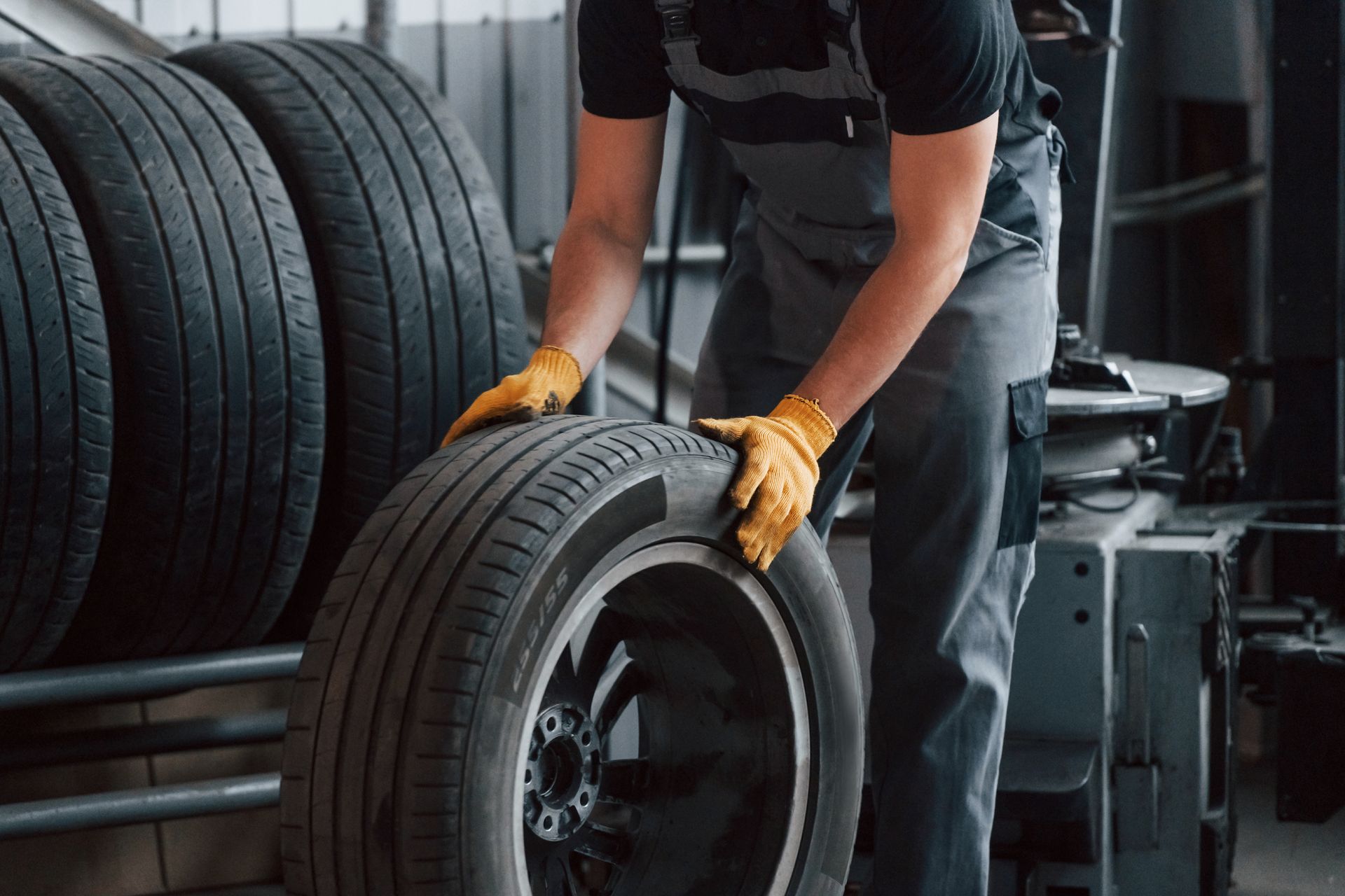 Mechanic in overalls and gloves holding a tire in a garage, tires stacked in background.