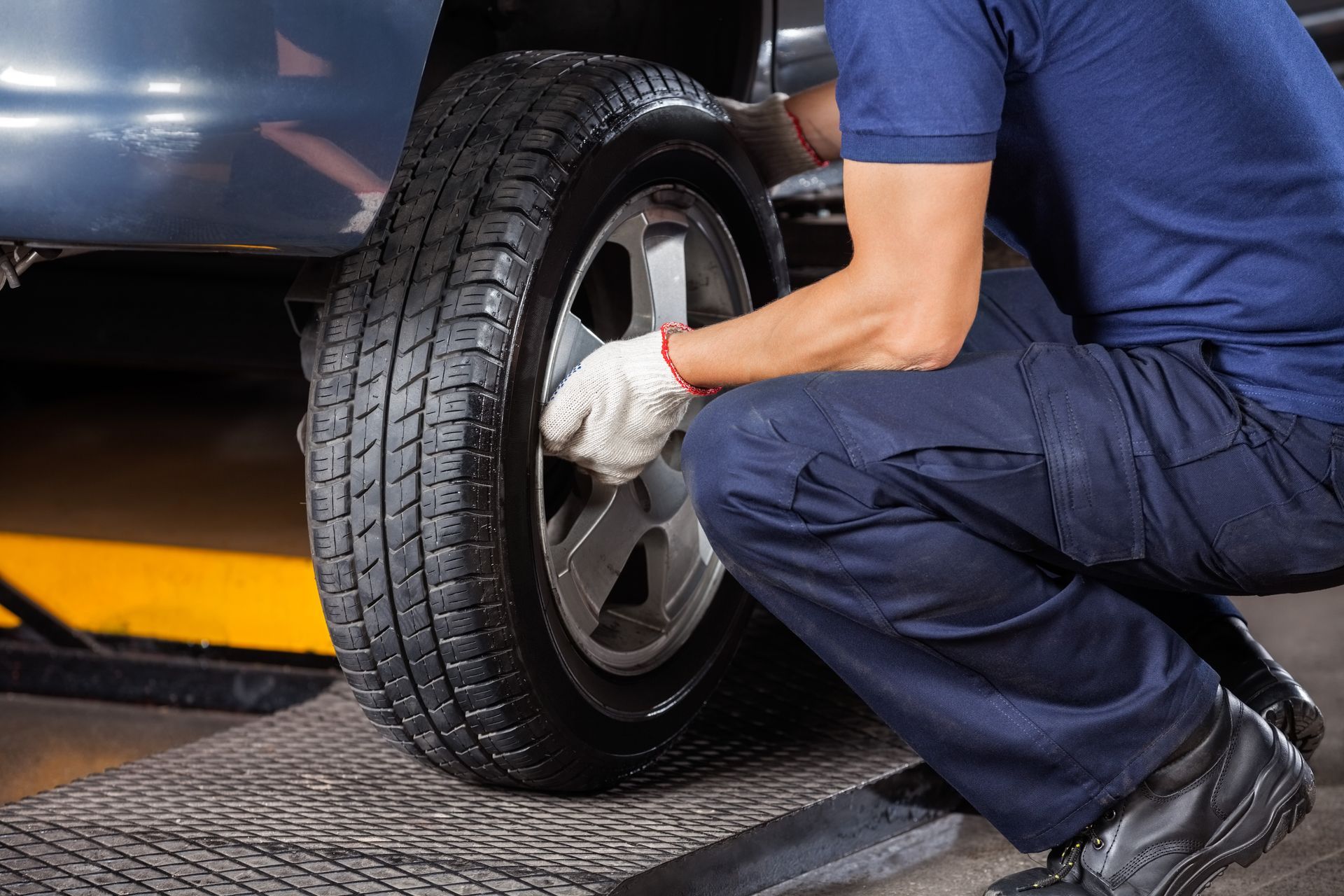 Mechanic in blue uniform changing a car tire on a ramp, wearing gloves.