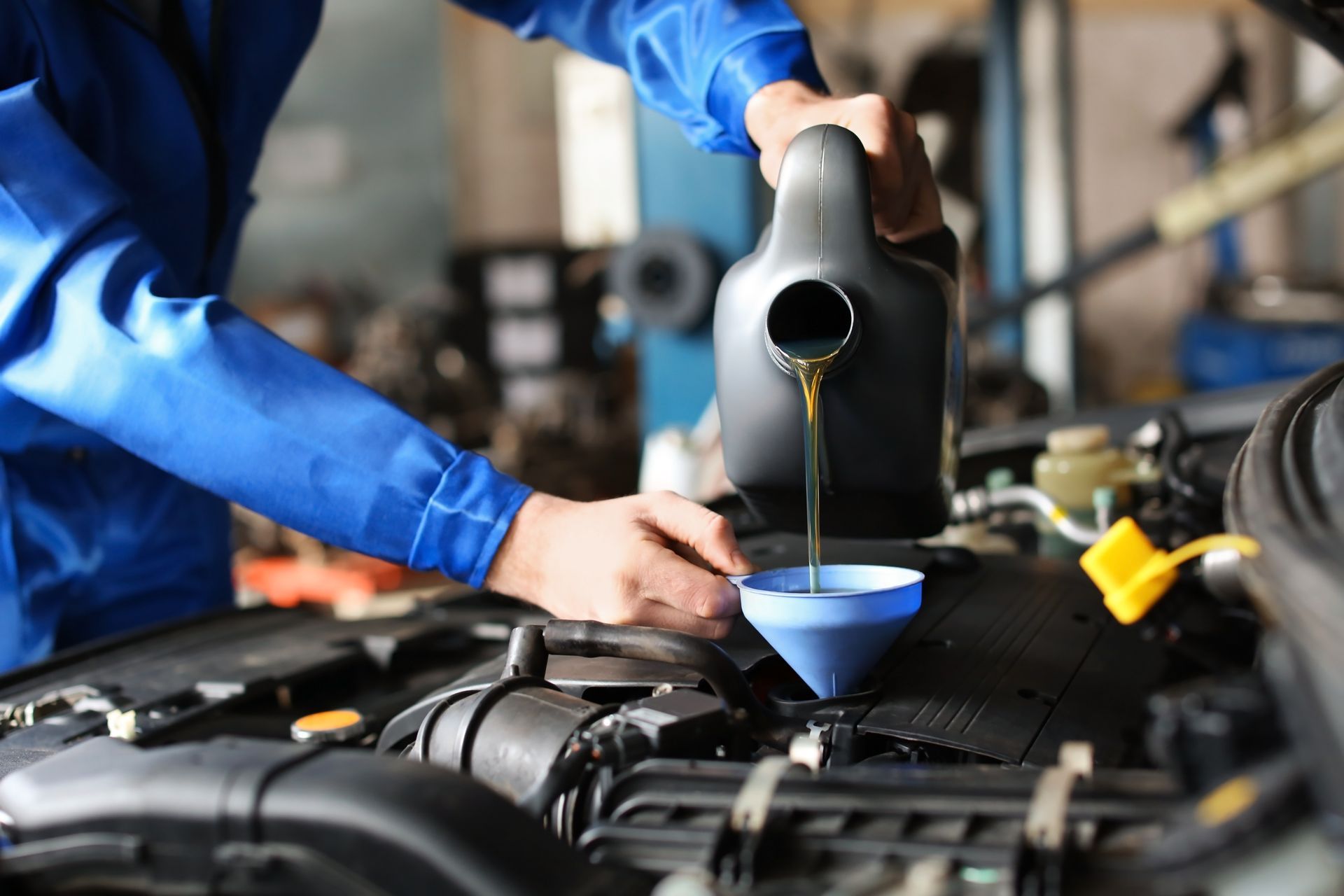 A mechanic in a blue uniform pours motor oil from a black container into a car engine using a blue funnel.