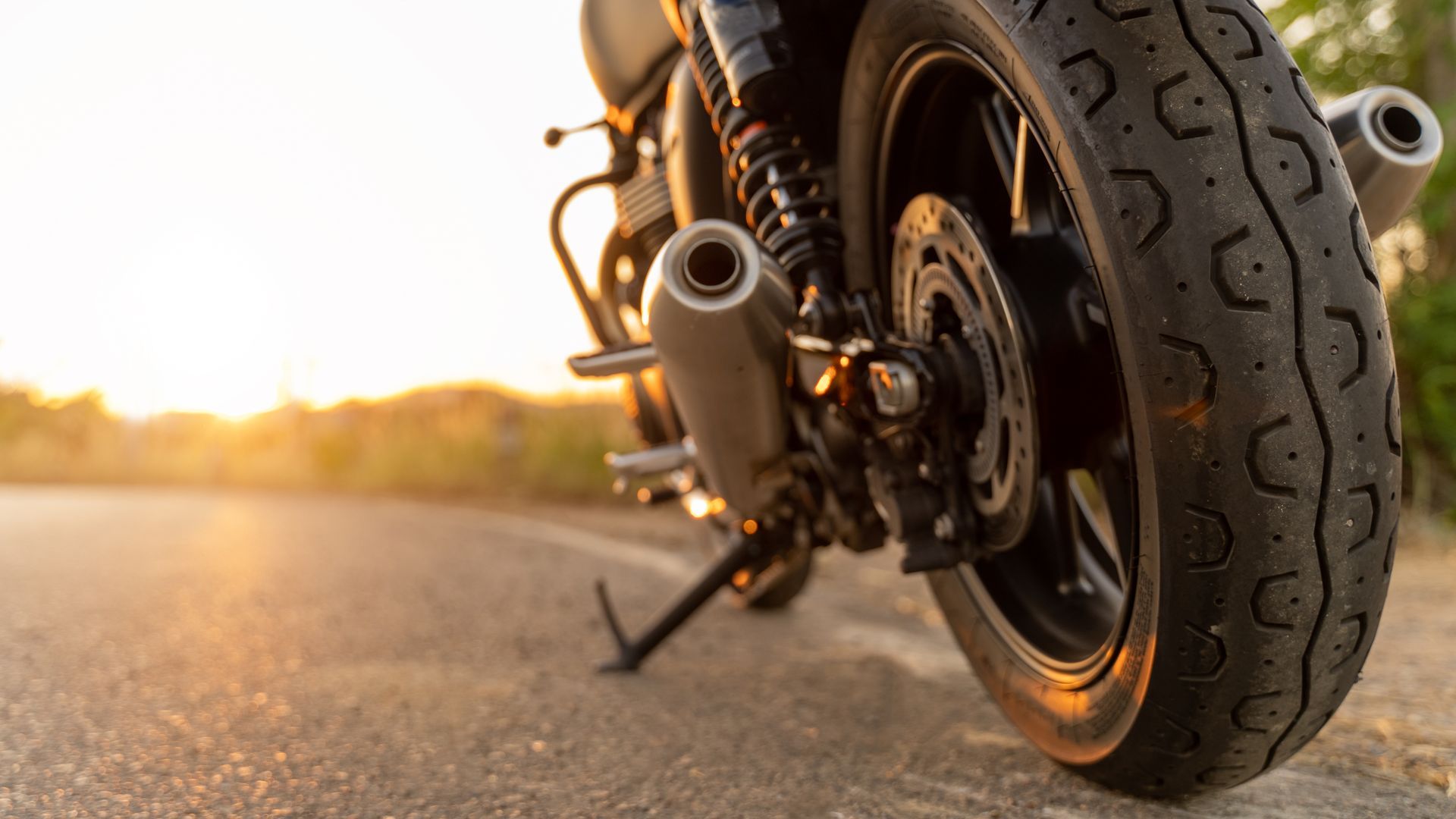 Close-up of a motorcycle rear wheel and exhaust on a sunlit road at sunset