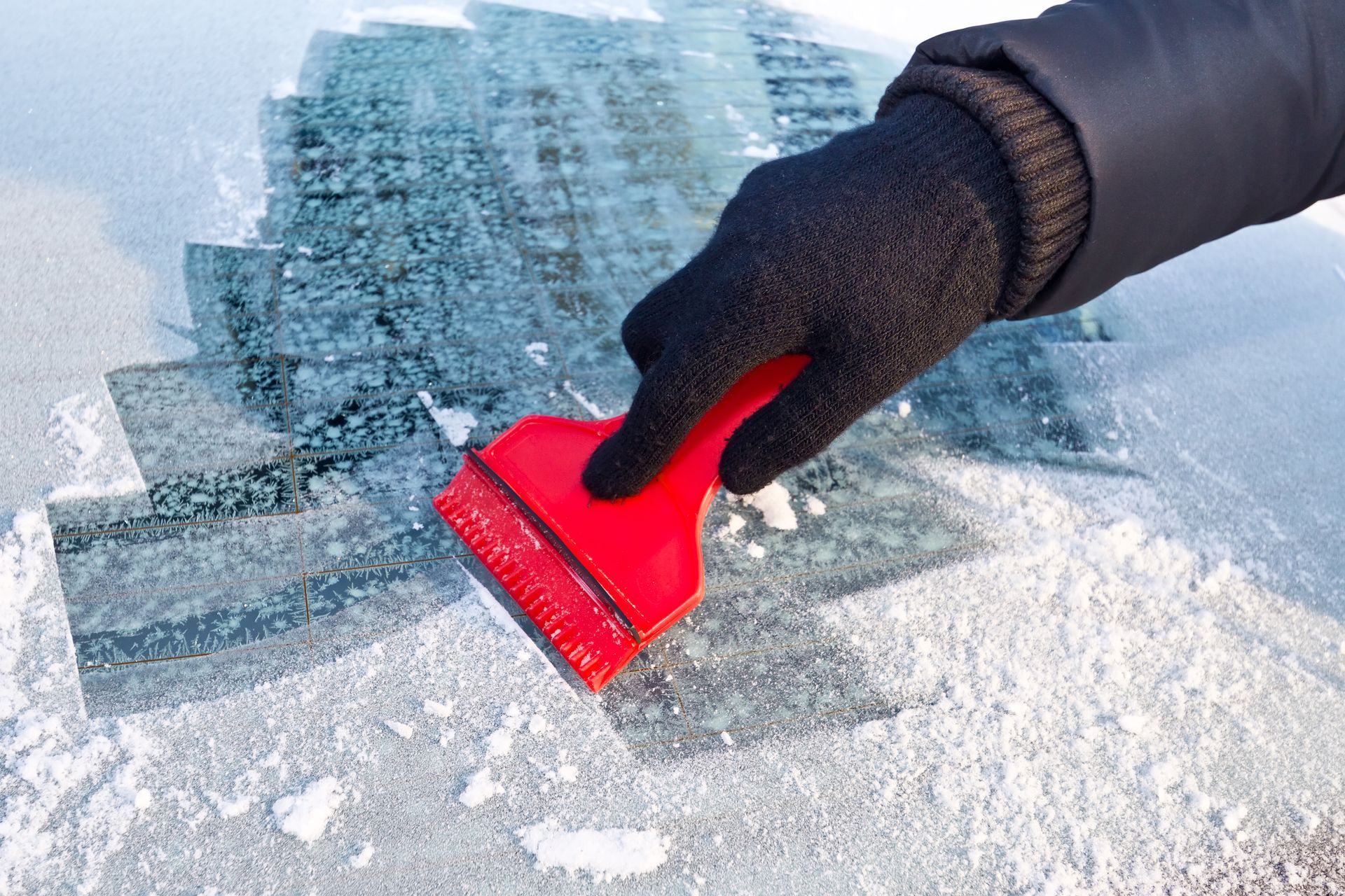 Gloved hand using a red scraper to clear ice from a car windshield.