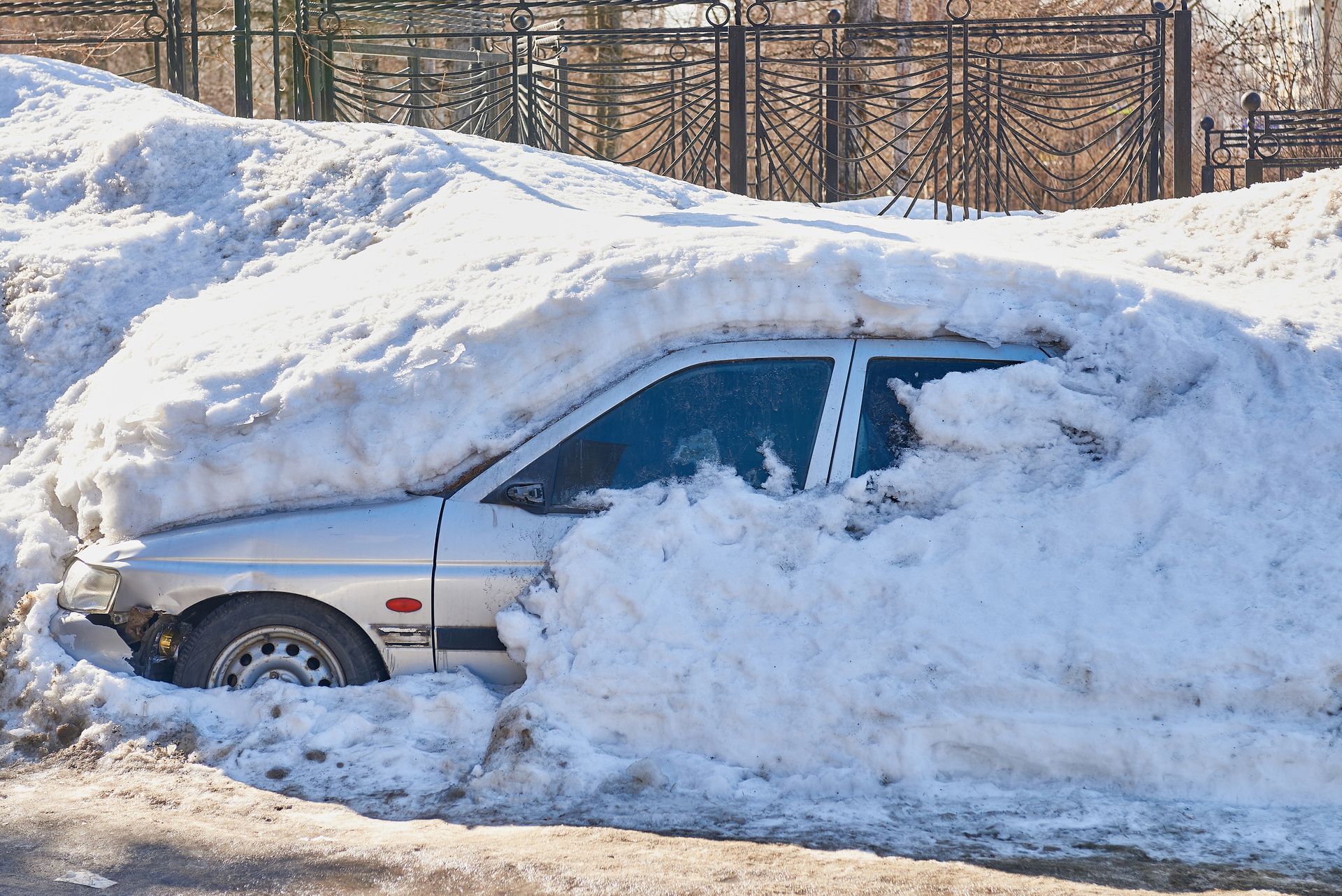 Silver car buried in a massive pile of snow on a road.