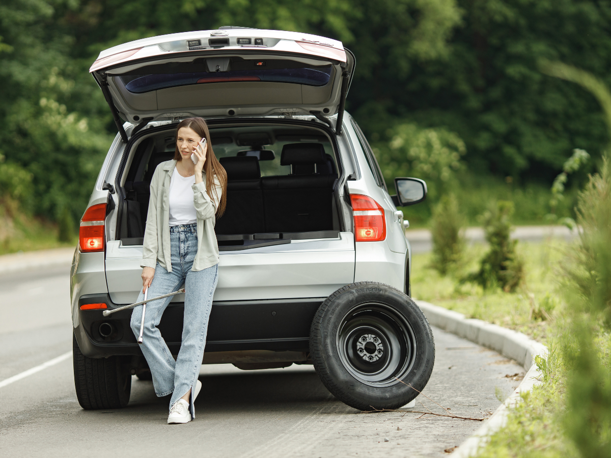 A woman is leaning on the back of a car with a flat tire and talking on a cell phone.