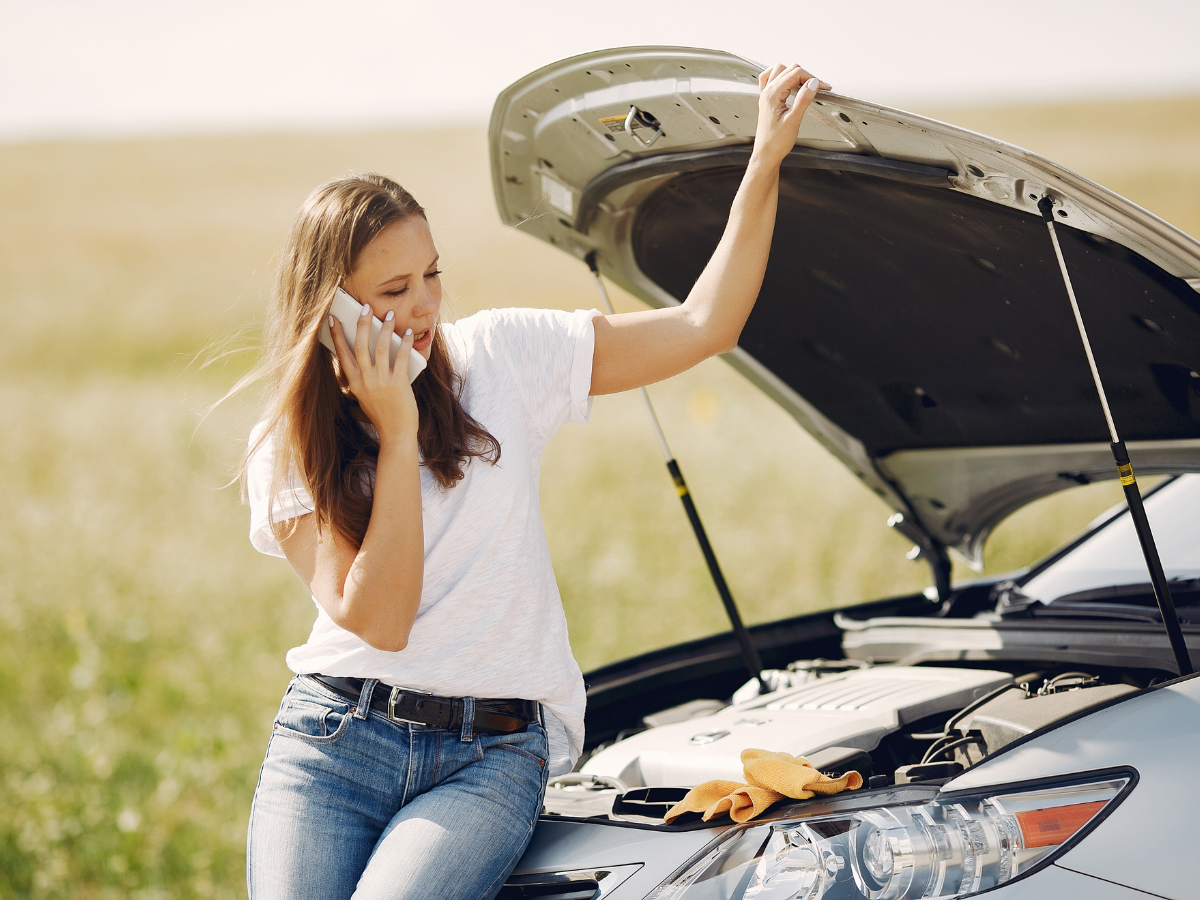 A woman is sitting on the hood of a broken down car talking on a cell phone.