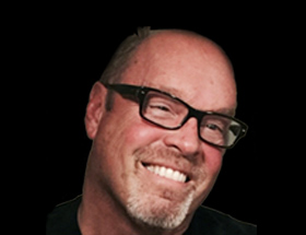 A person with a shaved head and facial hair, wearing black-framed glasses, smiles against a plain black background.