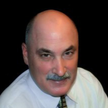 A headshot of a bald man with a mustache, wearing a white collared shirt and patterned tie against a black background.