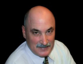 A person with a mustache, wearing a light-colored collared shirt and tie, against a solid black background.