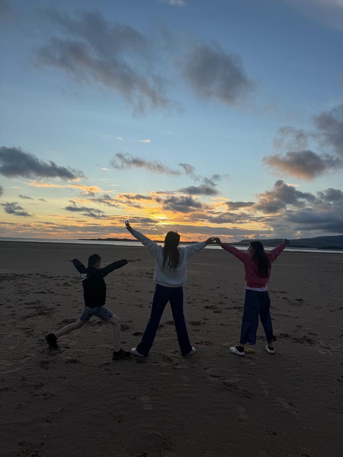 Three people stand on a beach at sunset, arms outstretched, silhouetted against a cloudy sky.