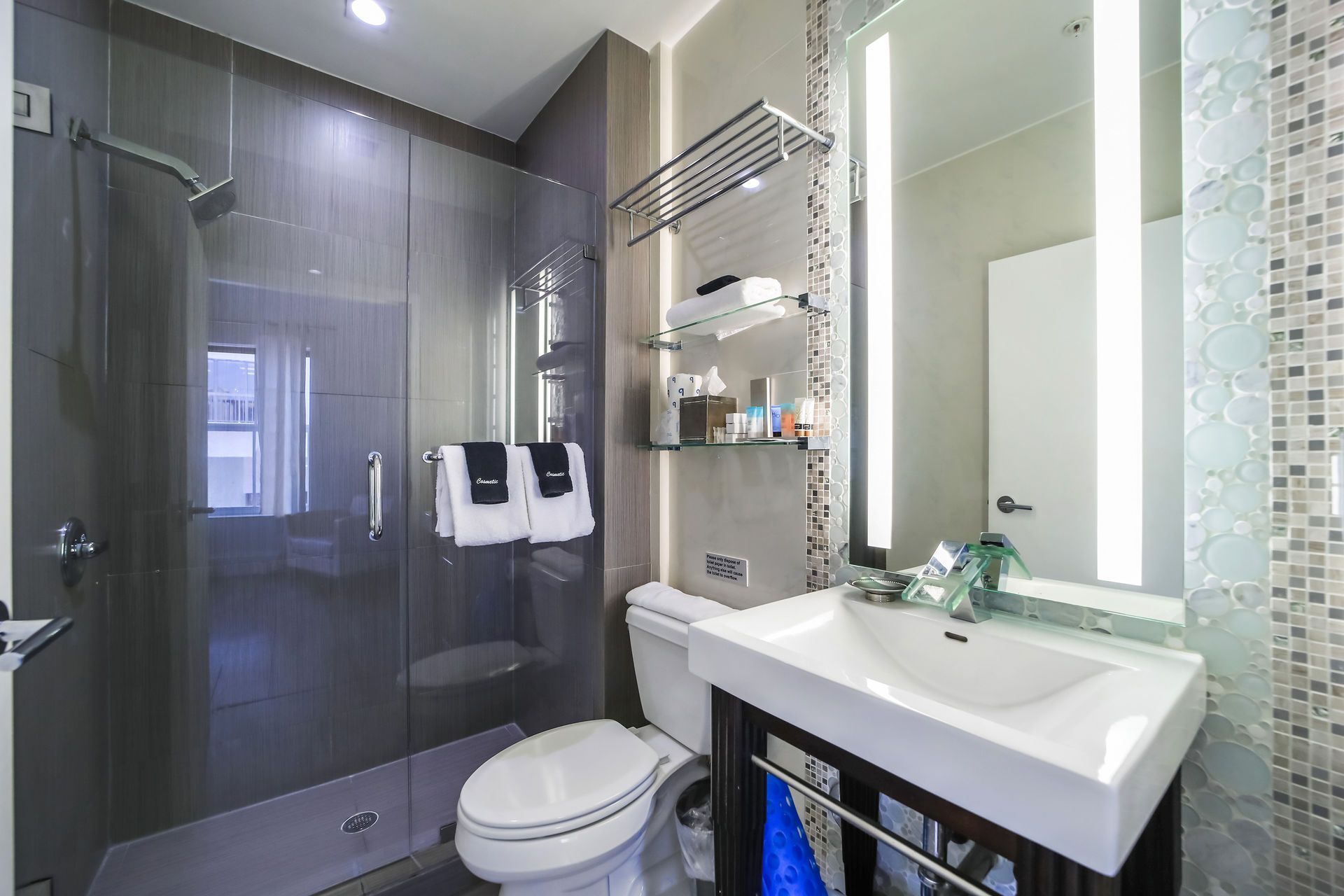 Modern bathroom with a shower, toilet, and sink. Gray walls, glass shower door, and a lit-up mirror.