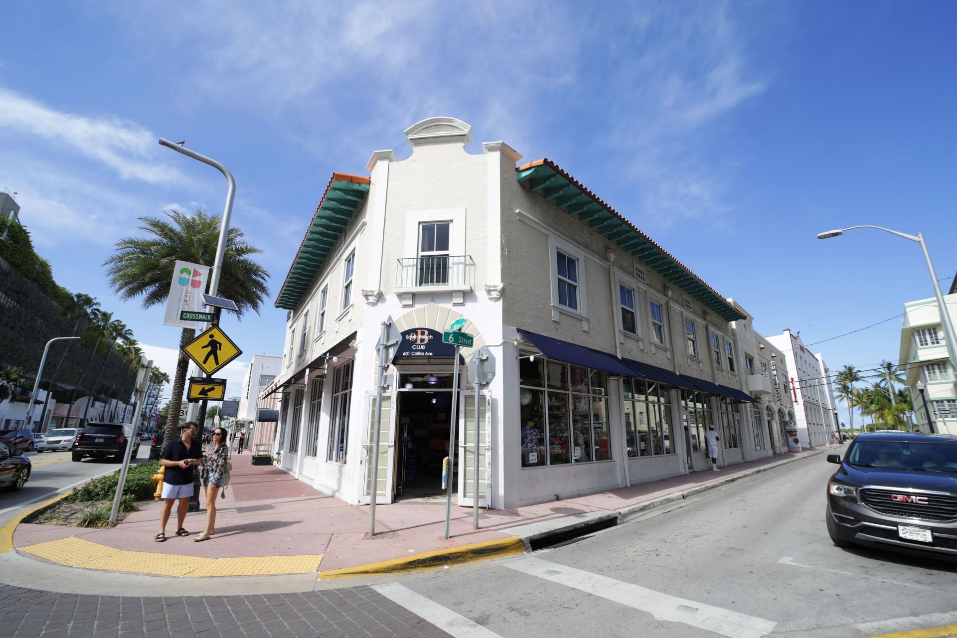 Corner building with shops, pedestrians on sidewalk, cars on street, blue sky.