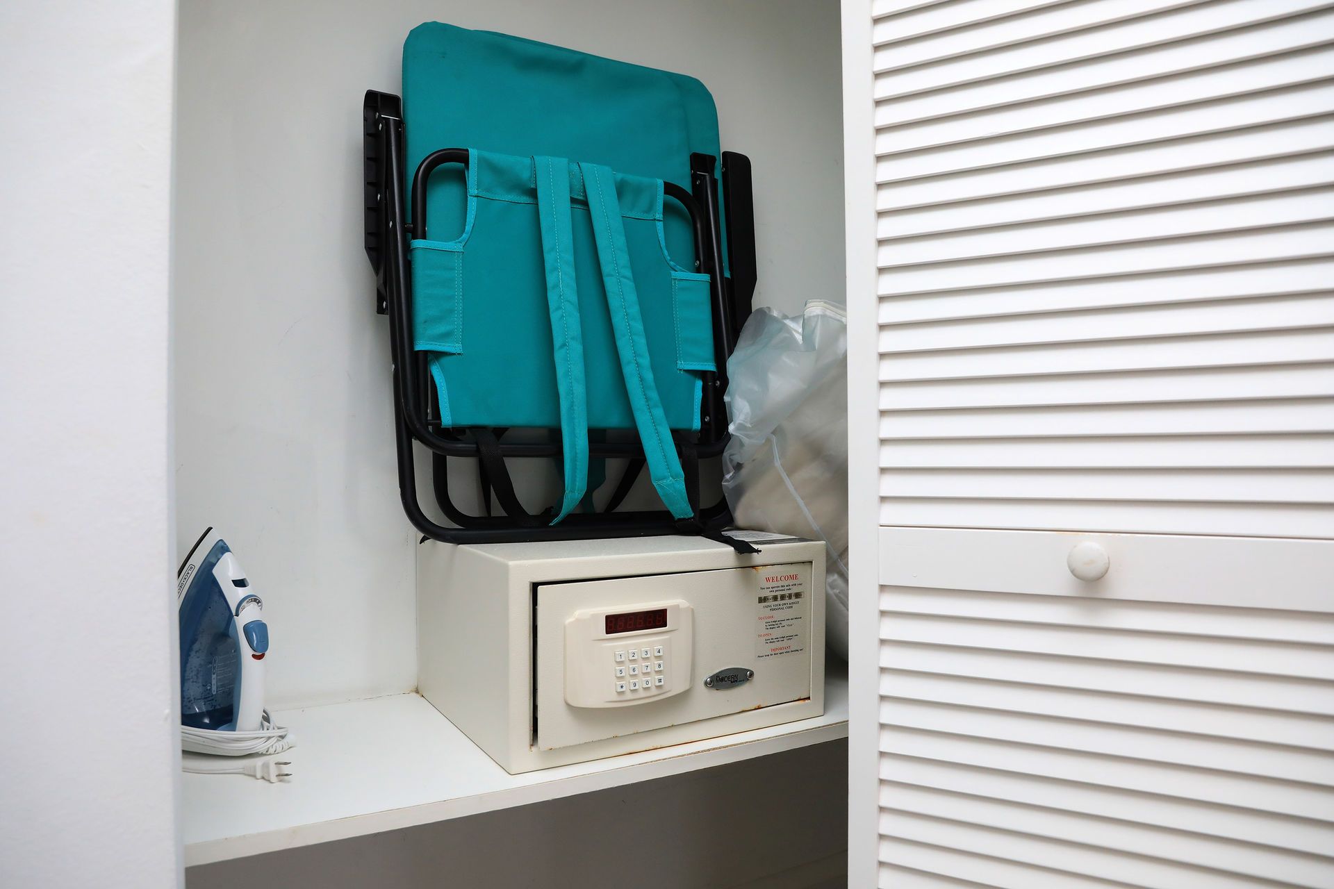 A teal folding chair sits atop a safe in a white closet next to an iron.