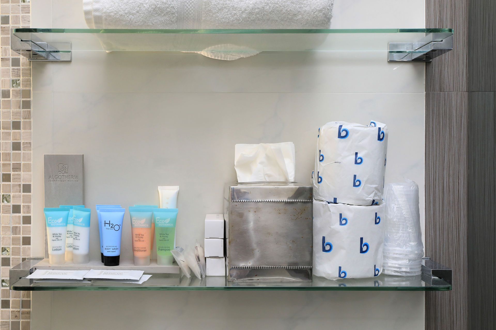 Bathroom shelf with toiletries: towels, tissue box, toilet paper, cups, and travel-sized toiletries.