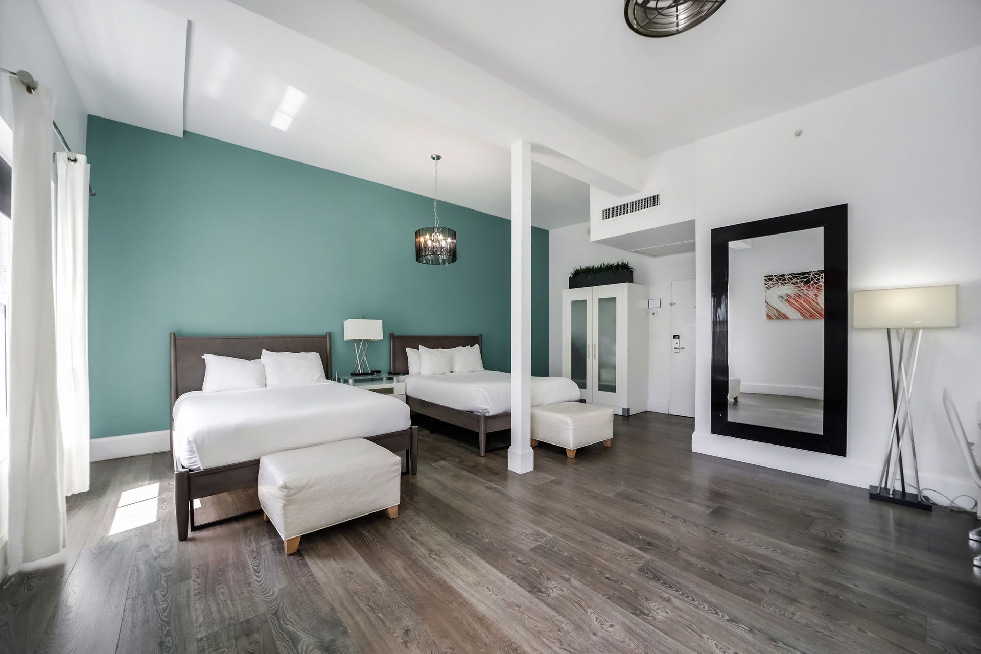 Spacious hotel room with two beds, large mirror, teal wall, wood floor, and natural light.