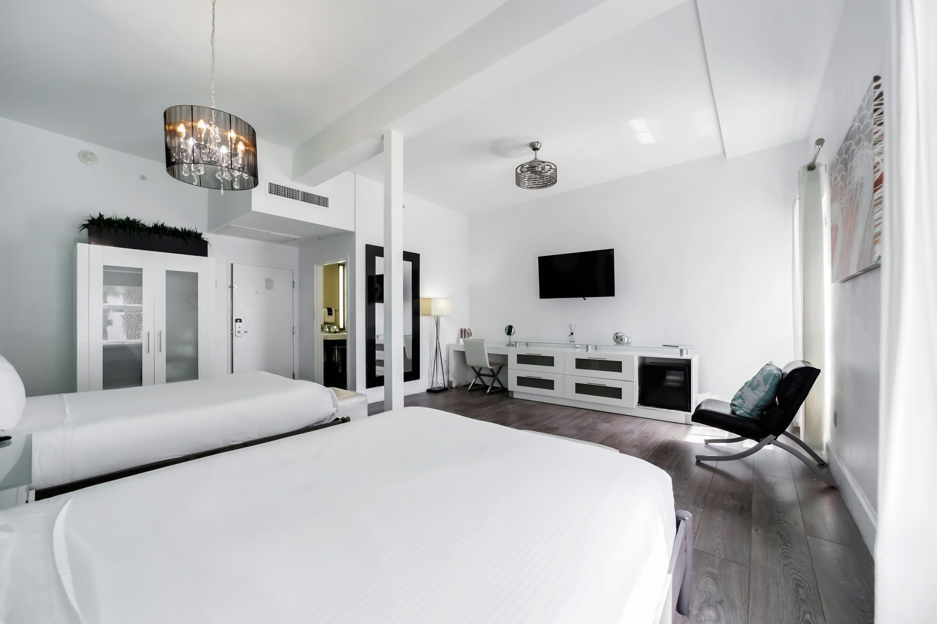 Bright white hotel room with two beds, TV, and black chair.