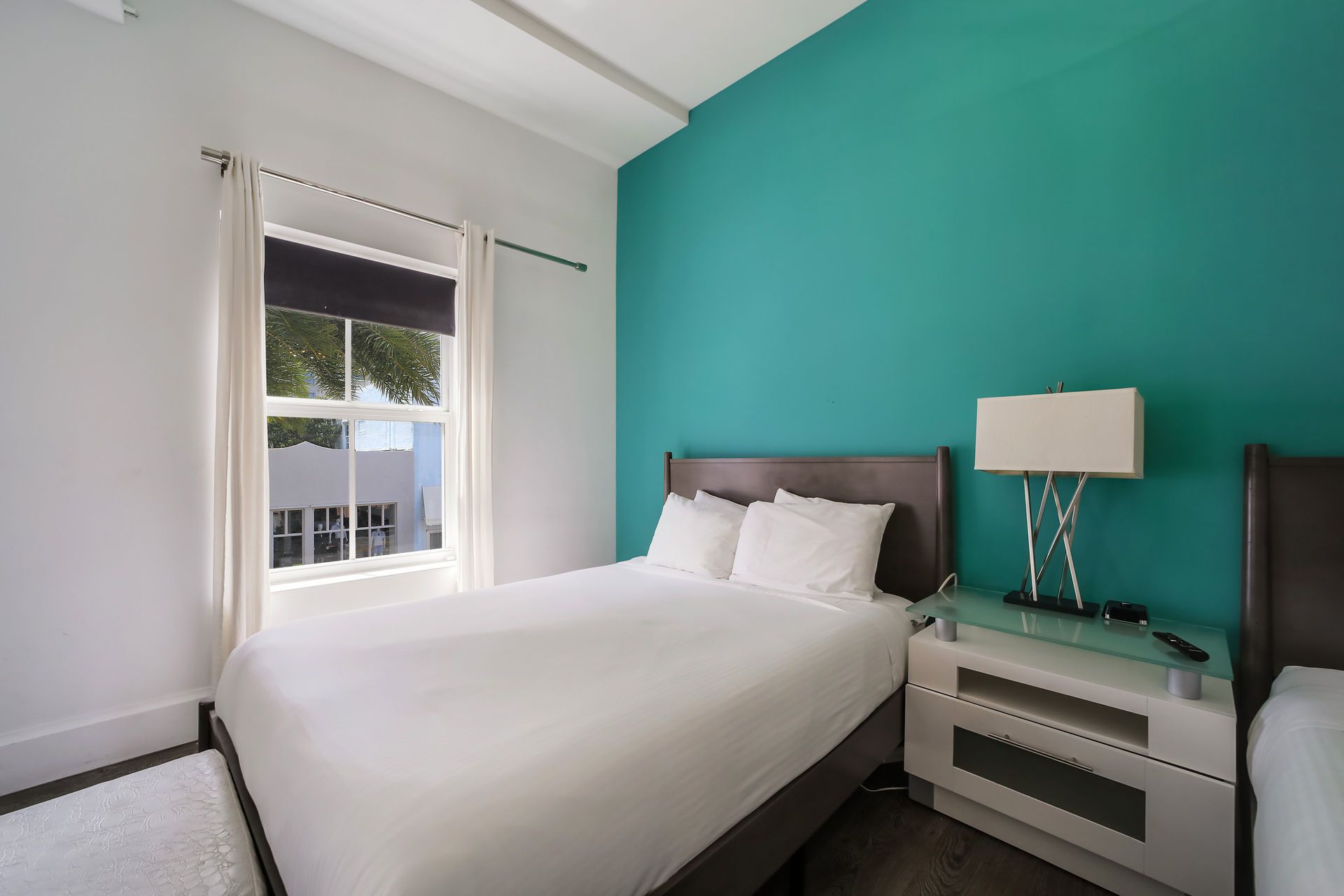Bedroom with two beds, turquoise accent wall, window with white trim, and a bedside table.