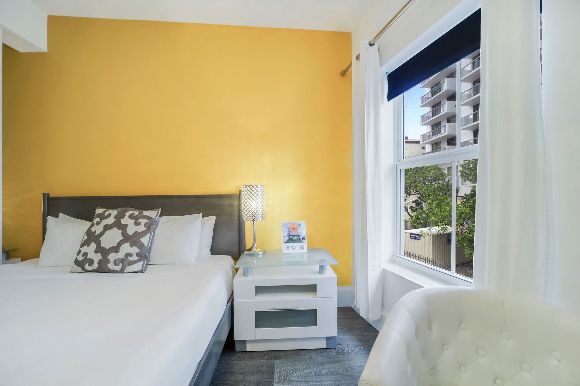 Yellow-walled hotel room with a bed, nightstand, window, and a white chair.